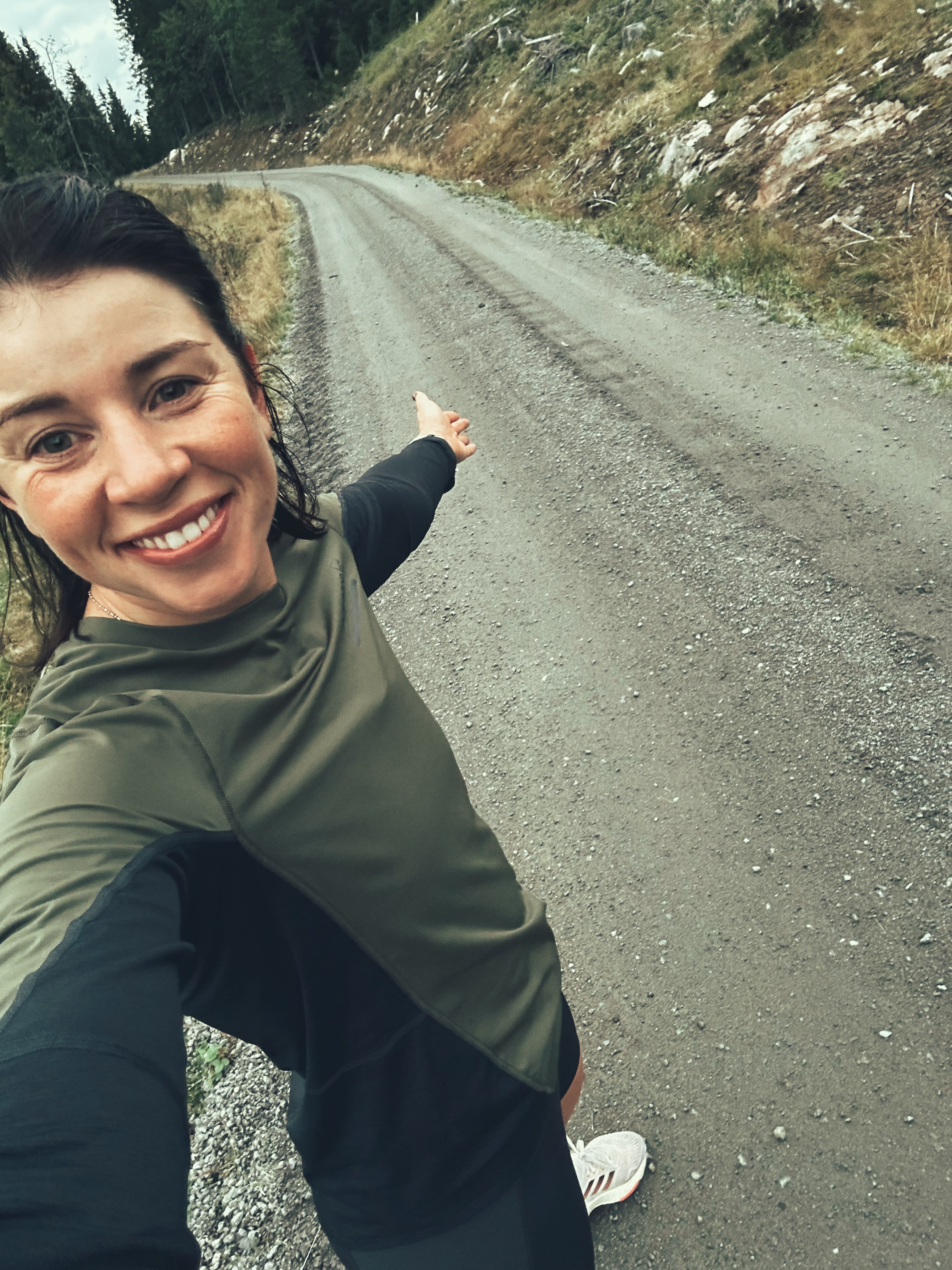 A woman in a Norrøna shirt and black leggings smiles and points toward a gravel road.