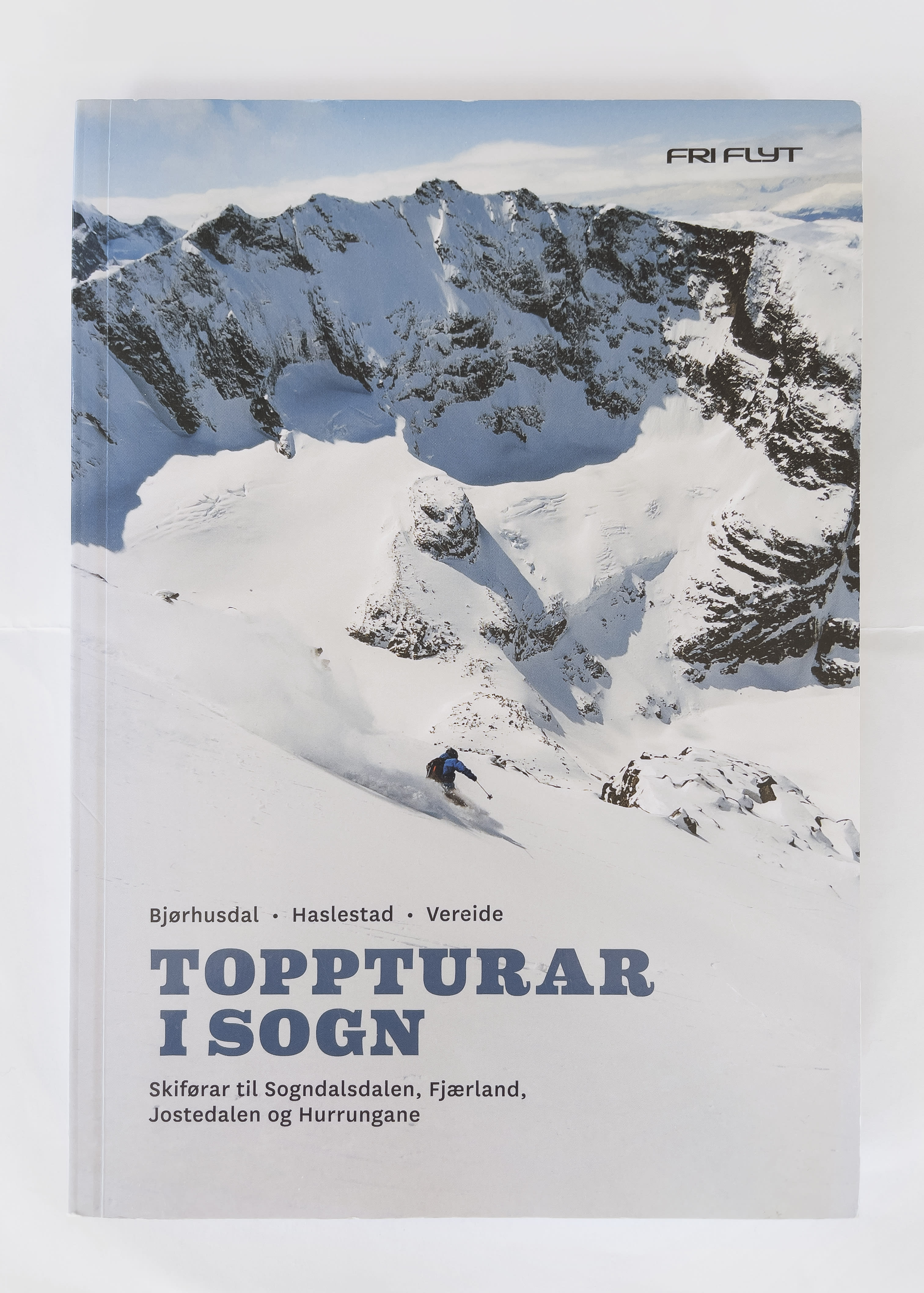 A skier wearing a Norrøna jacket descends a snowy mountain slope. The image is a book cover for "Toppturar i Sogn", a guide to skiing in the Sogn region of Norway. 
