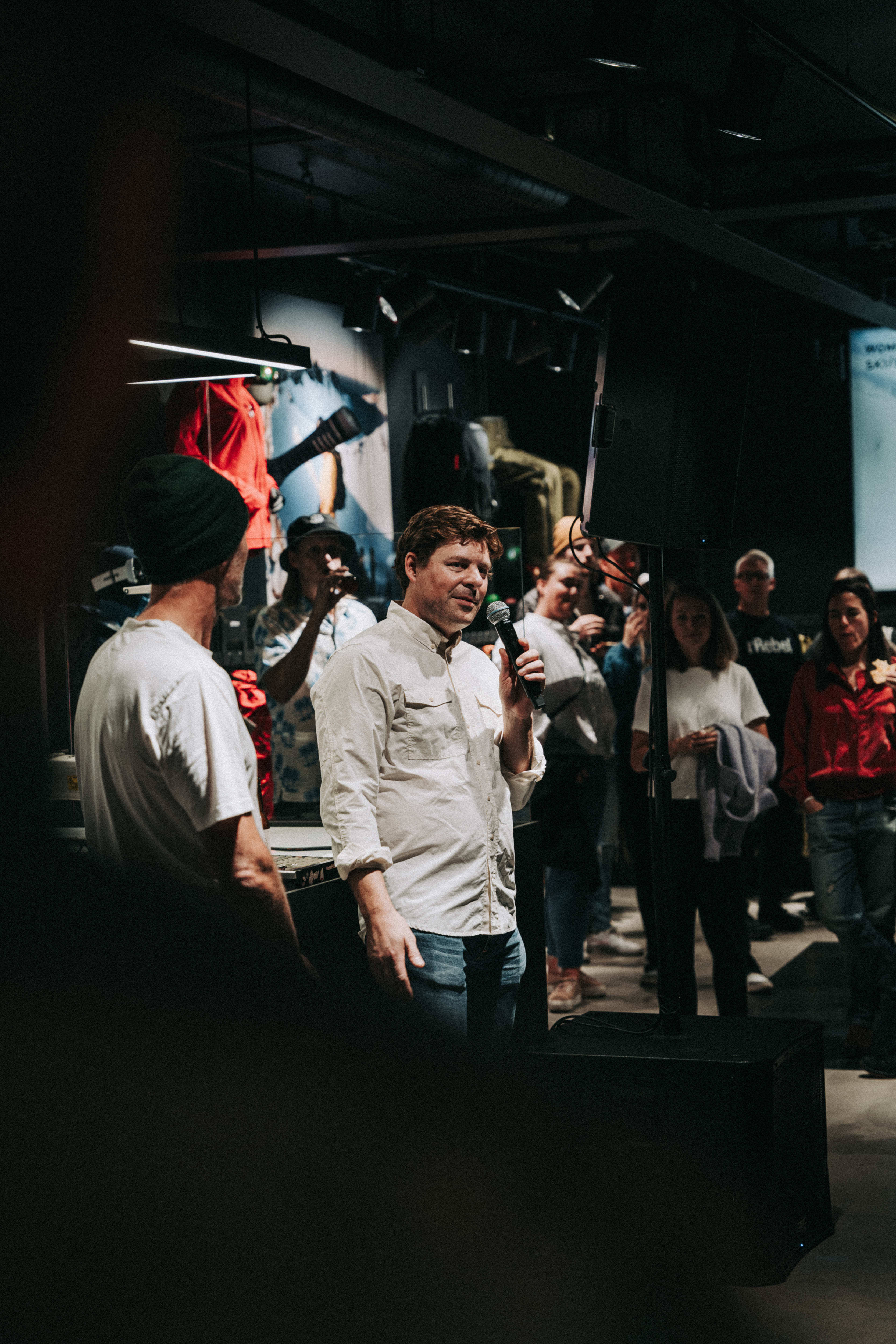 A man speaking into a microphone in a Norrøna store, surrounded by other people.  A red Norrøna jacket hangs on a rack in the background. 

