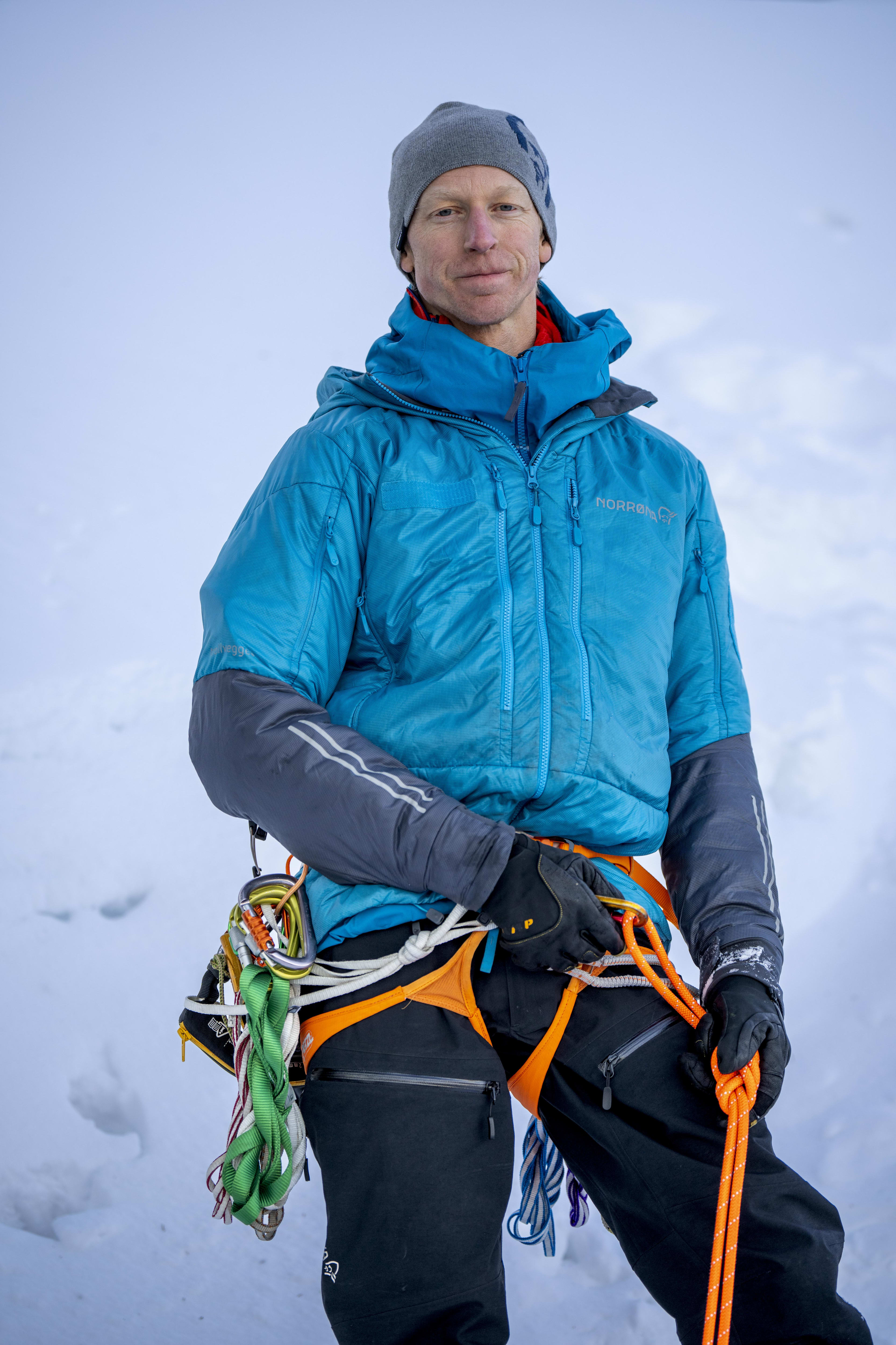 A man in a Norrøna jacket and pants stands in a snowy environment holding an orange climbing rope.