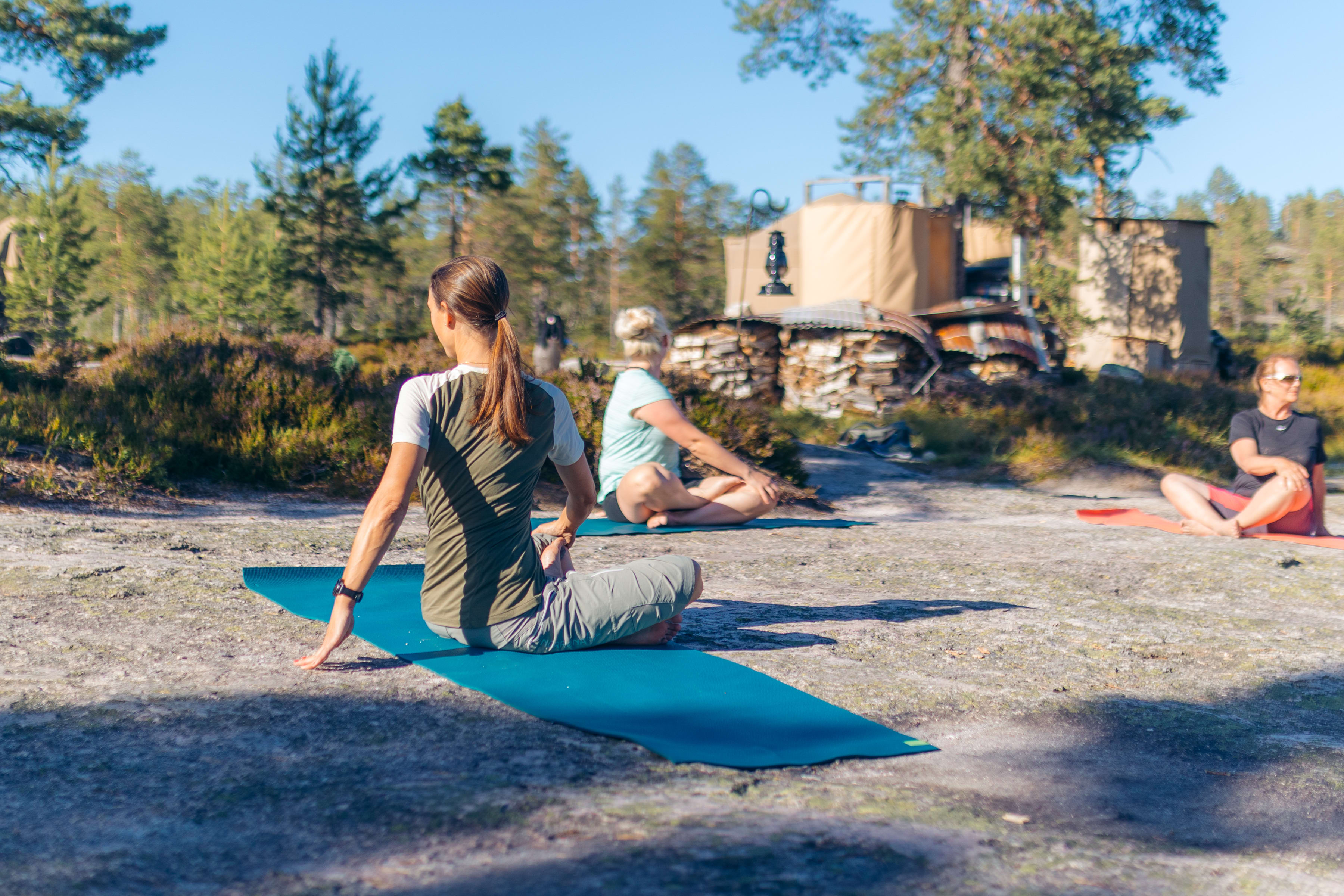A woman wearing a Norrøna shirt is sitting on a blue yoga mat in a yoga pose. Two other people are sitting on yoga mats in the background.