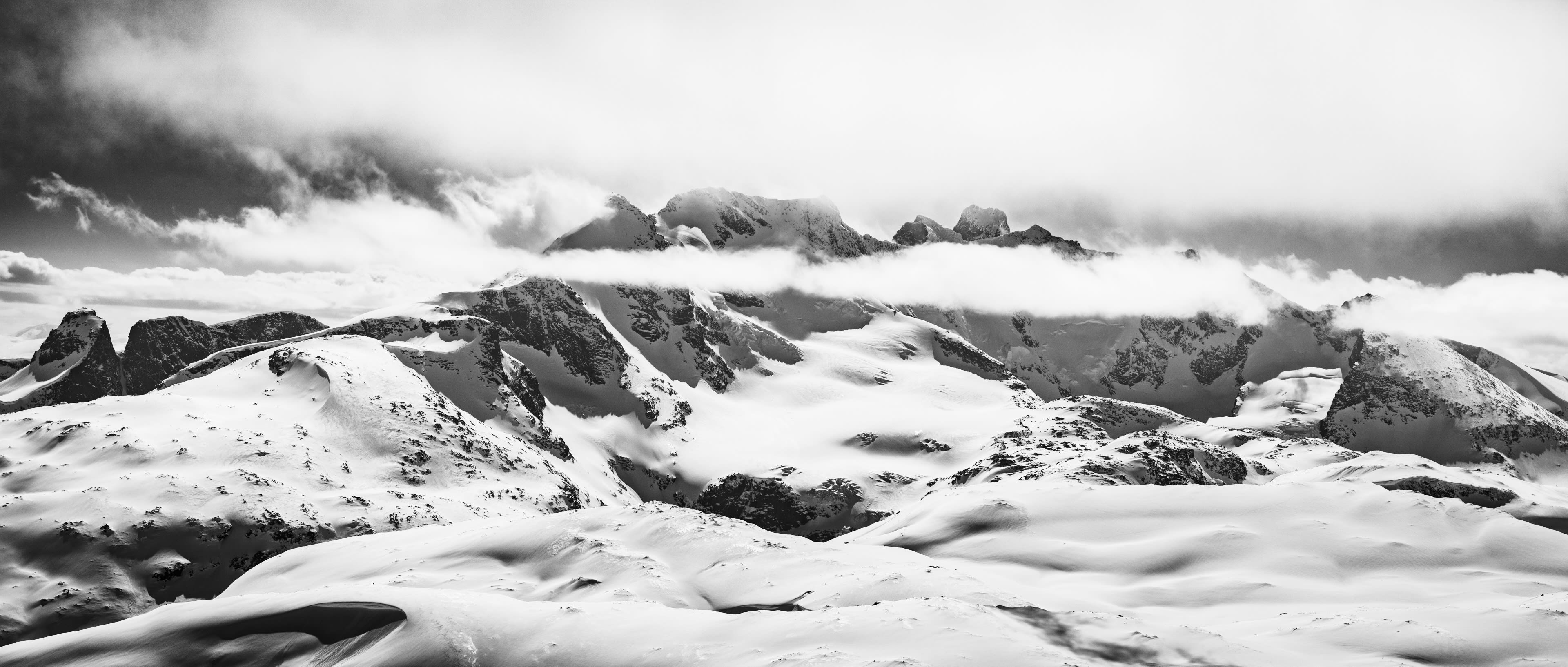 A black and white photograph of a mountain range covered in snow and clouds. The peaks are obscured by the clouds.