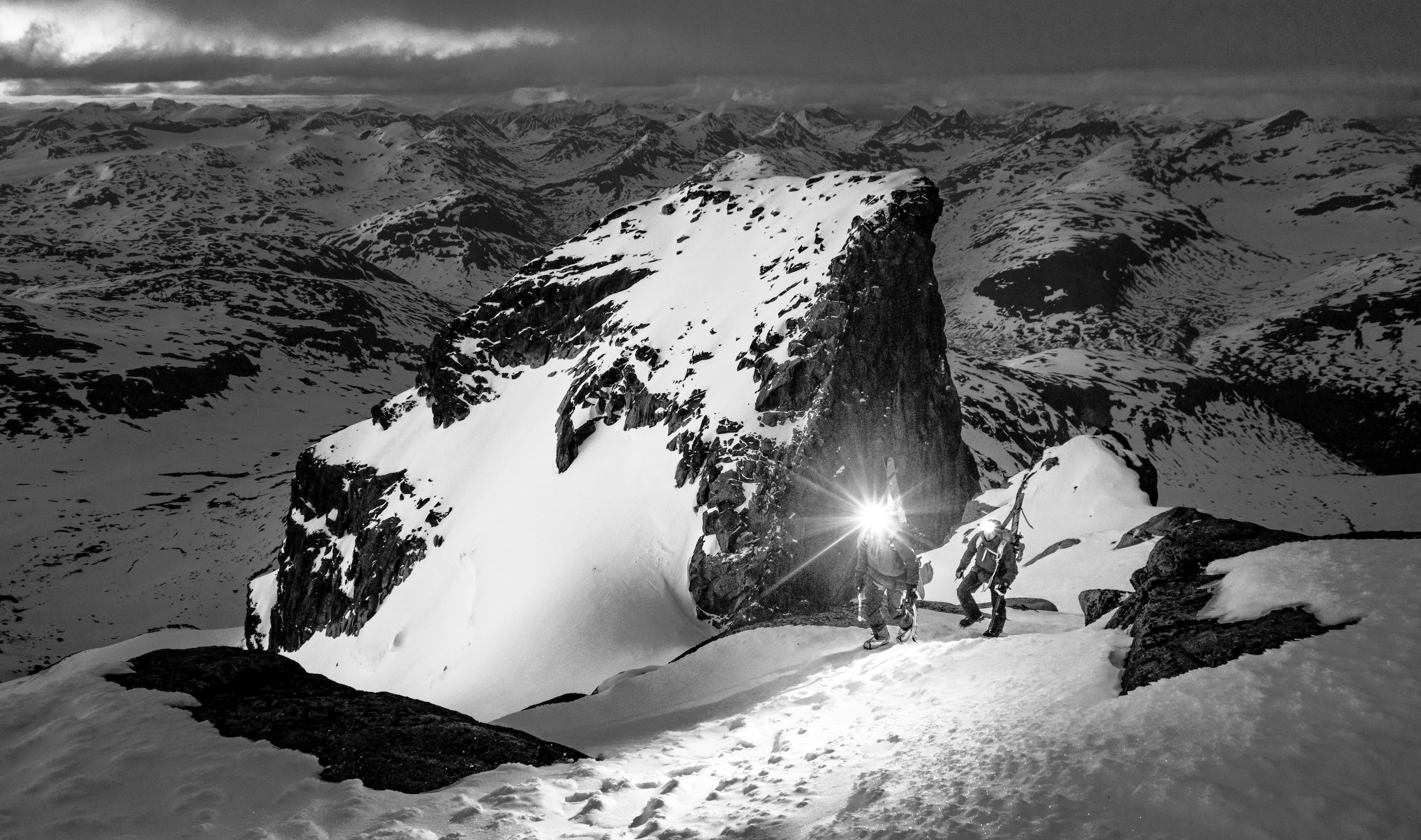 Two people are hiking through a snow-covered mountain range. They are wearing winter gear.  The person in the lead is holding a headlamp, which illuminates the snow-covered path.  The image is in black and white.