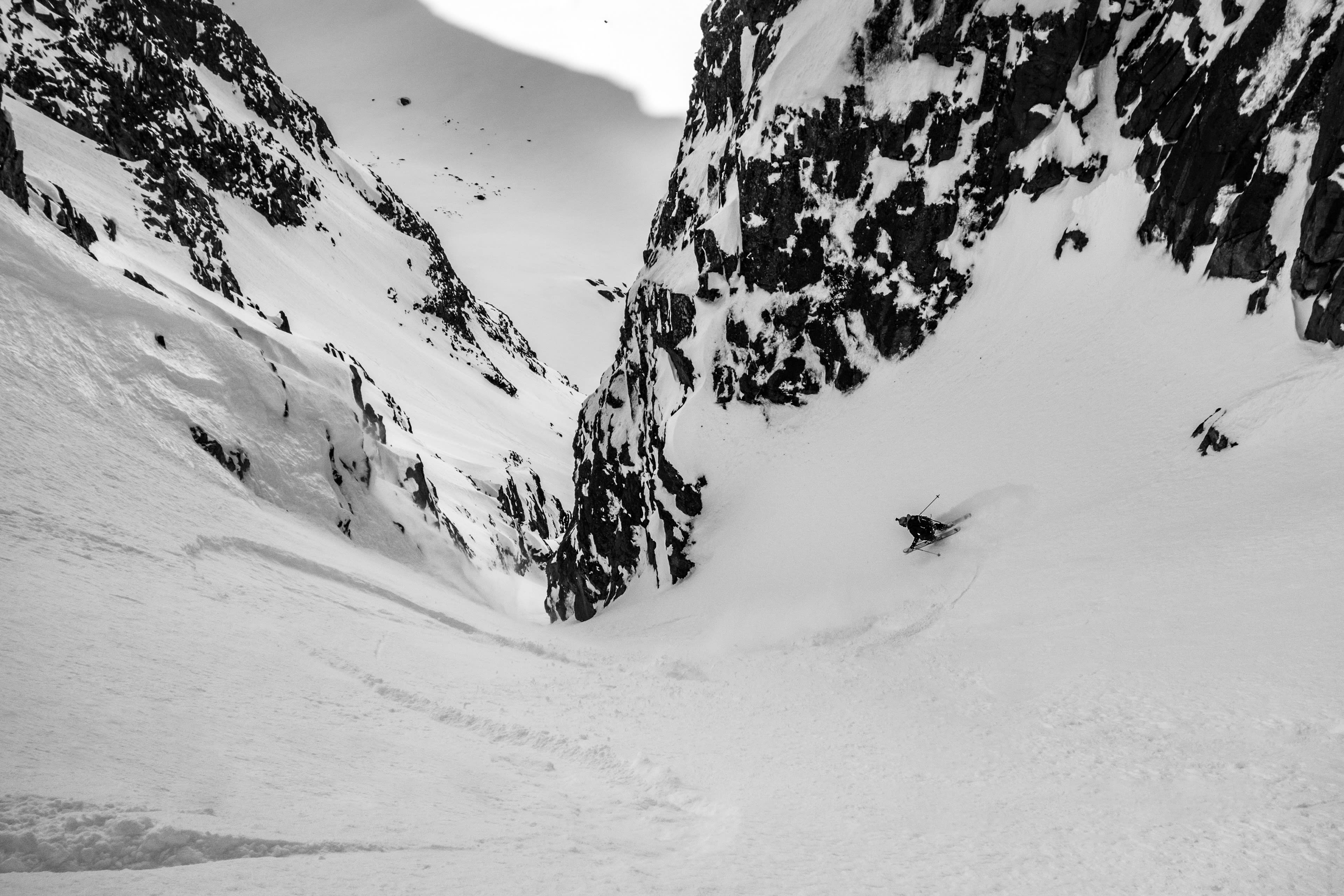 A person skis down a snow-covered mountain slope. The skier wears a black Norrøna jacket.  
