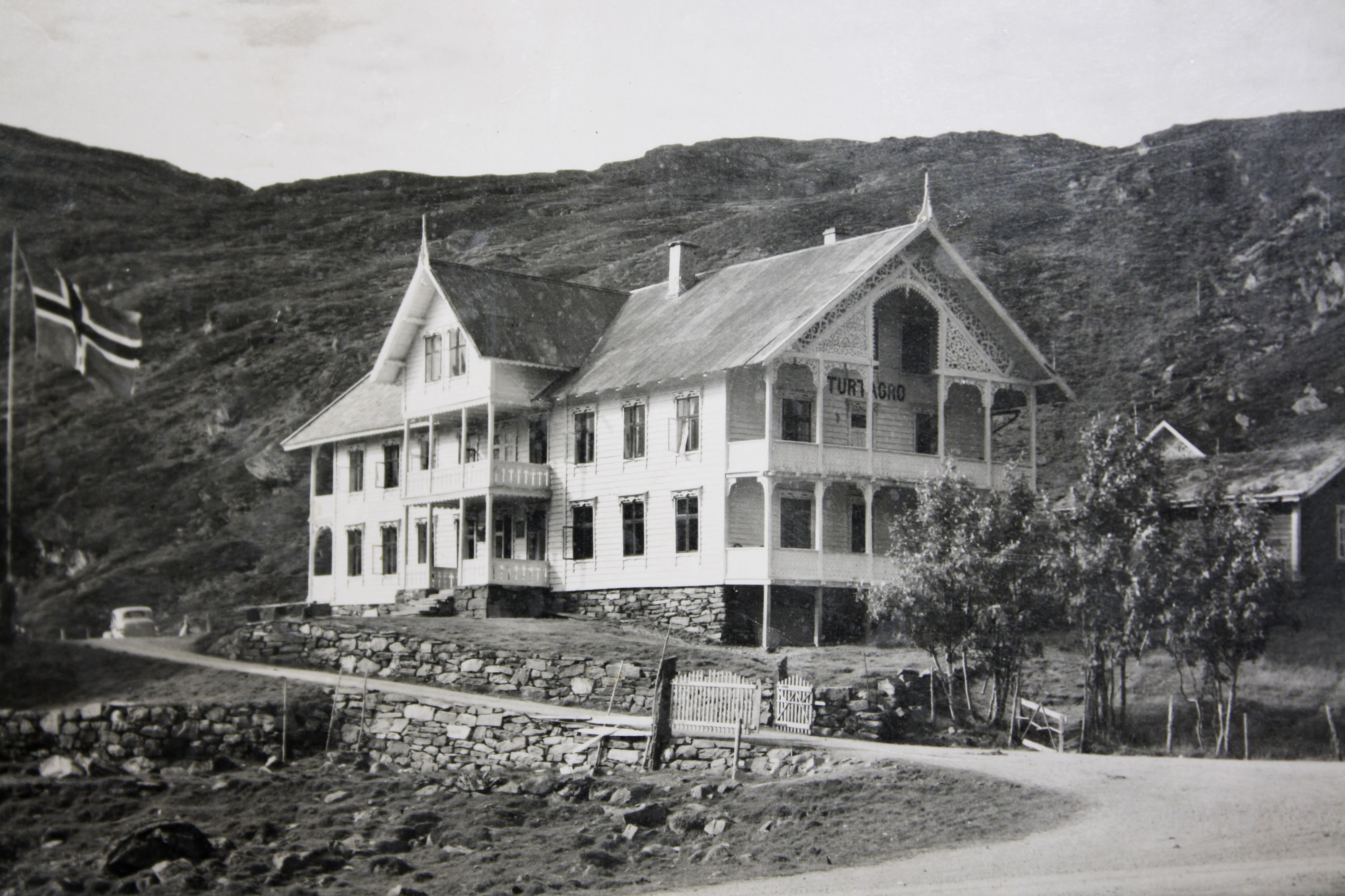 Black and white photo of a wooden house with a Norwegian flag in the background. The house has a sign above the door that says "Turtjågro".  A car and a stone wall are visible in the foreground.  The photo is likely from the early 20th century. 
