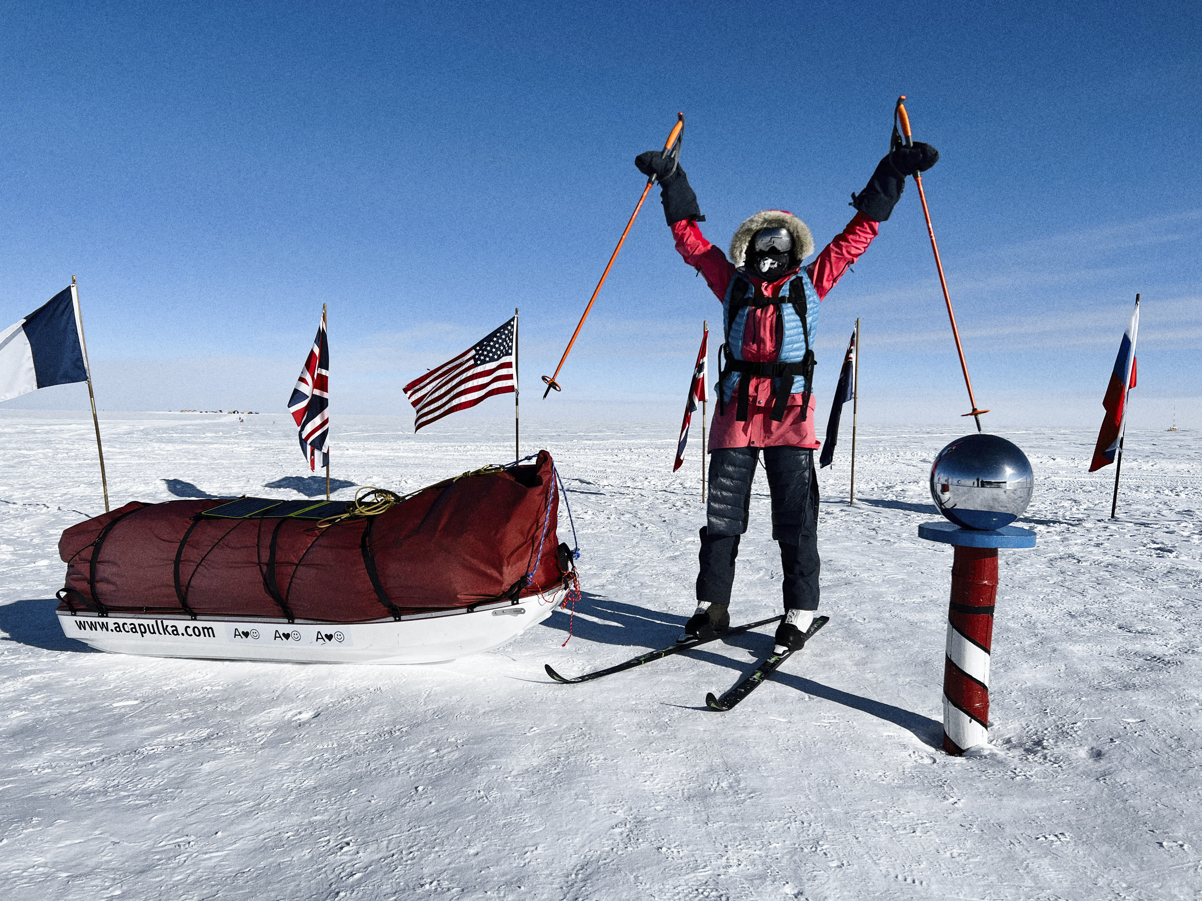 A person is standing in the snow with their arms raised holding ski poles, a red and white striped pole is beside them and a sled is behind them. There are flags in the distance.