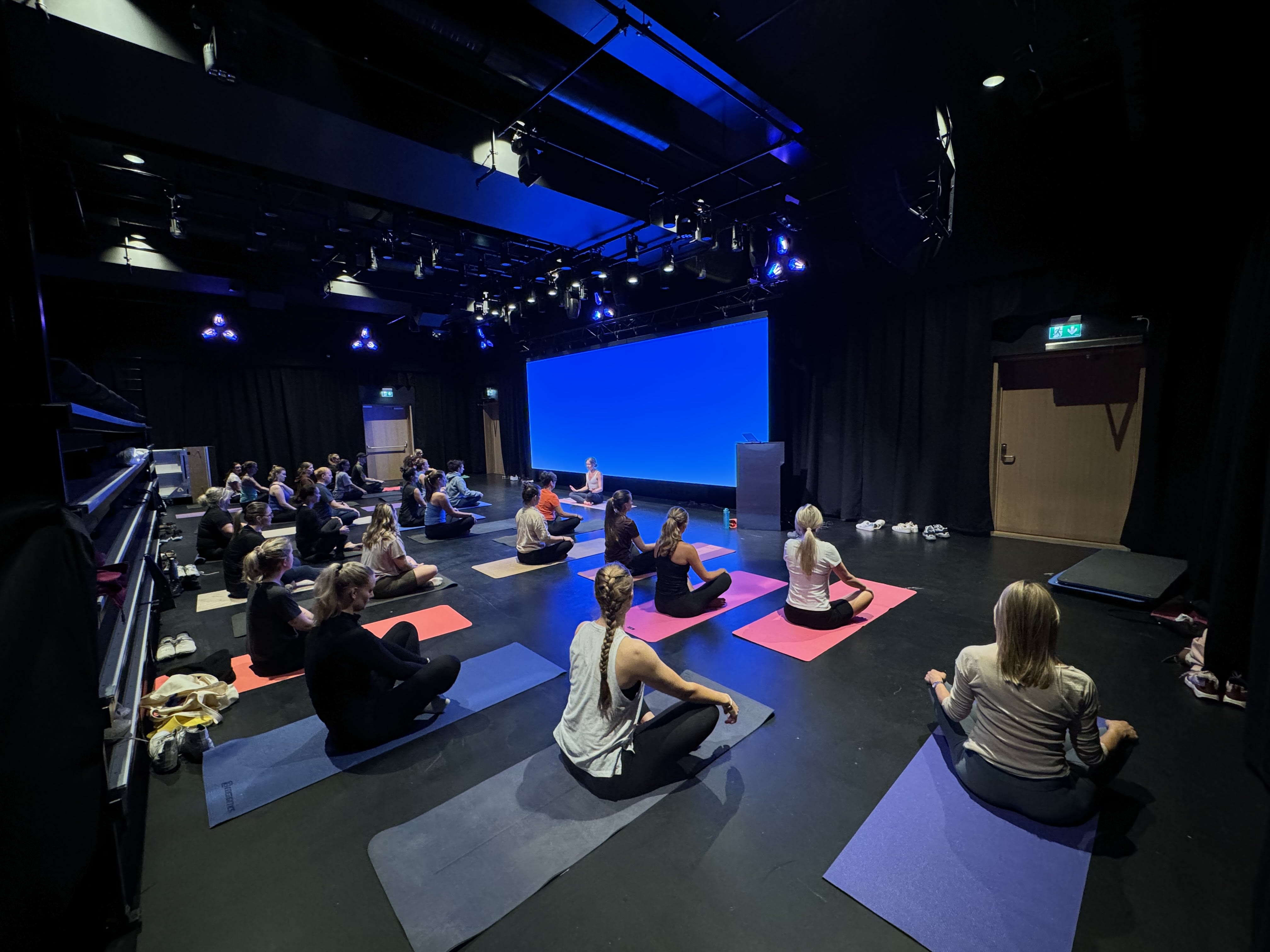 A group of people sitting in a yoga class. A large blue screen is behind them. Some are wearing Norrøna clothing.