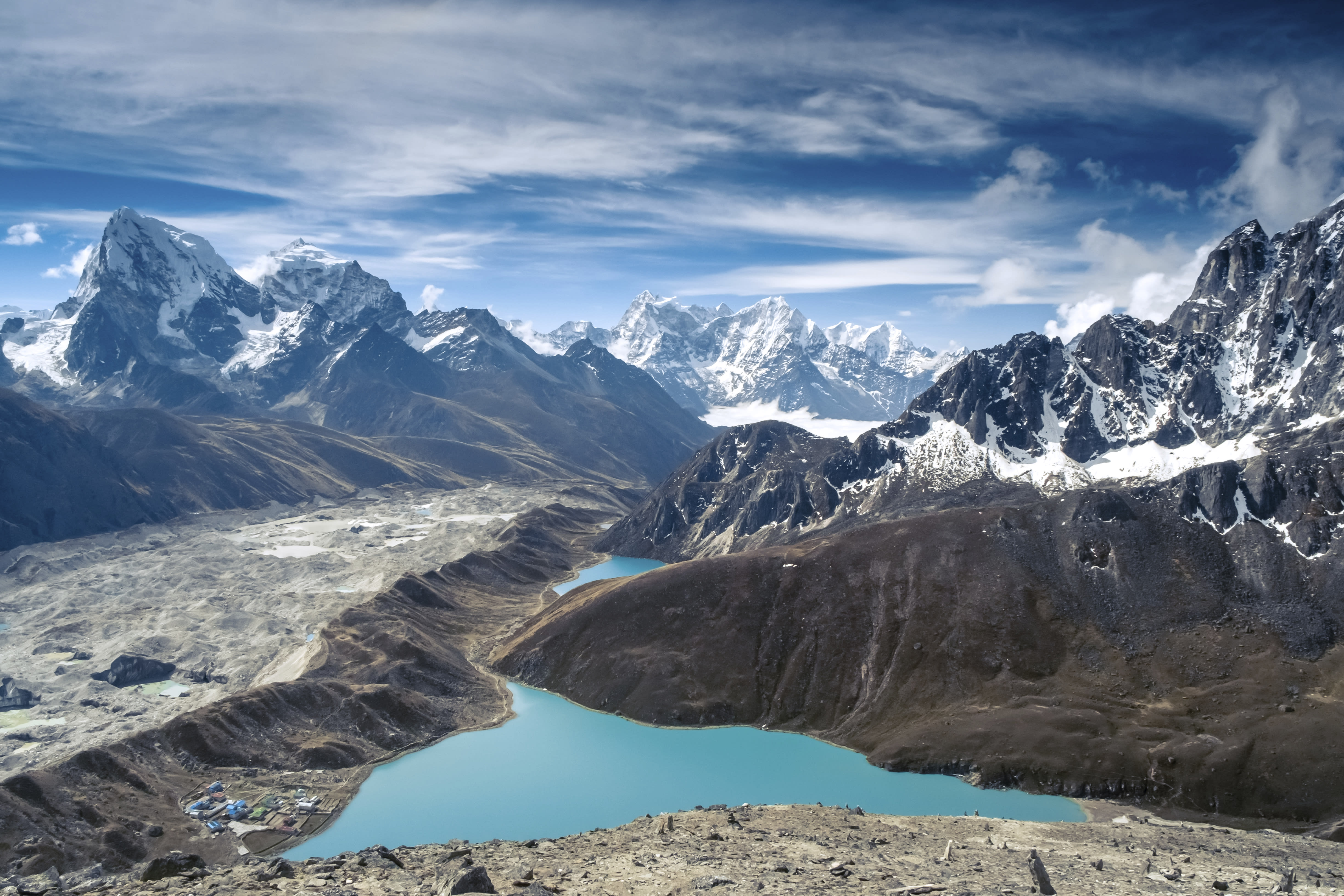 Scenic view of snow-capped mountains, blue lakes, and a small village under a blue and cloudy sky.
