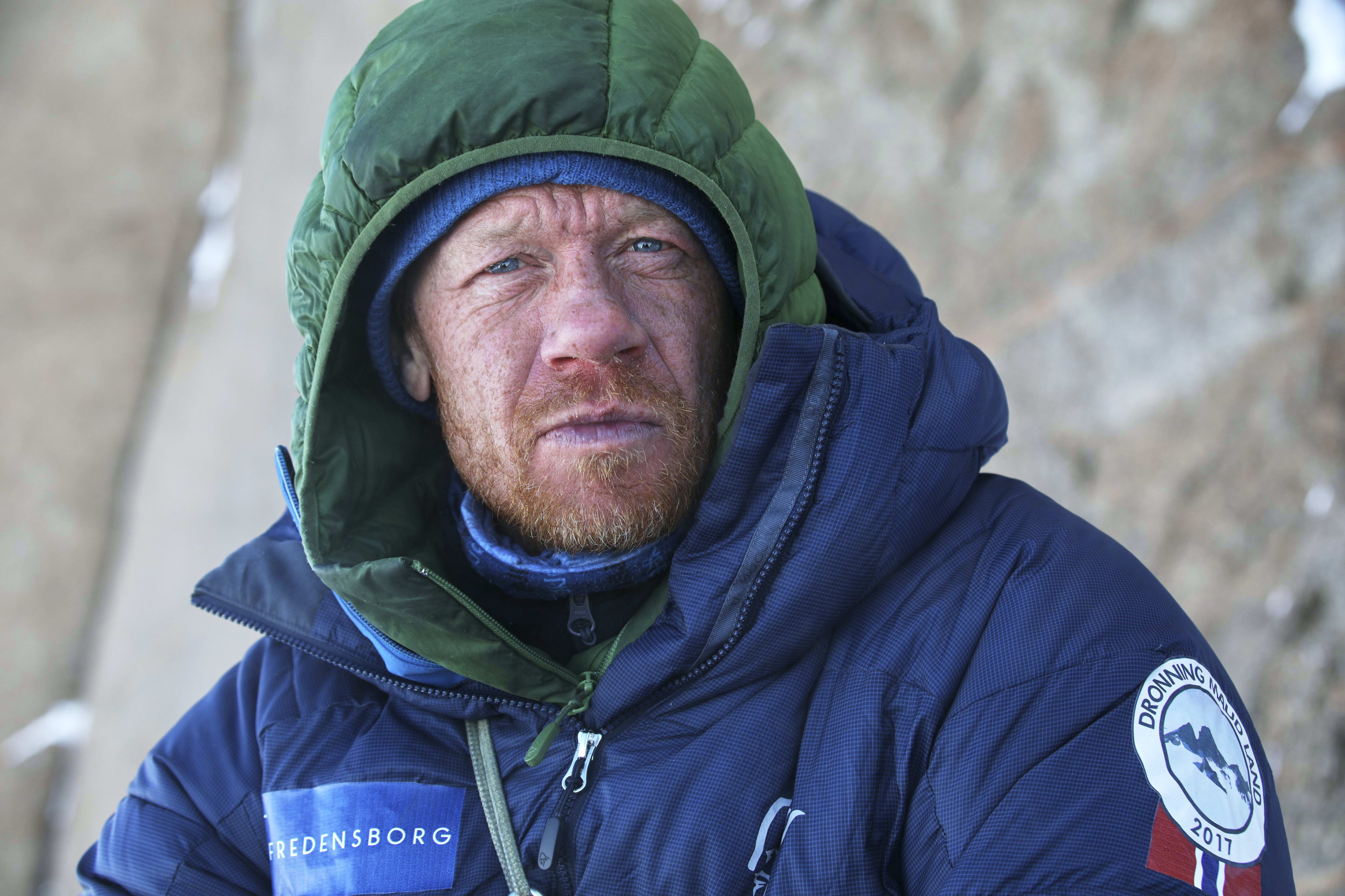 A man wearing a green and blue Norrøna jacket with a hood pulled up, looking to the right. He has a beard and blue eyes.