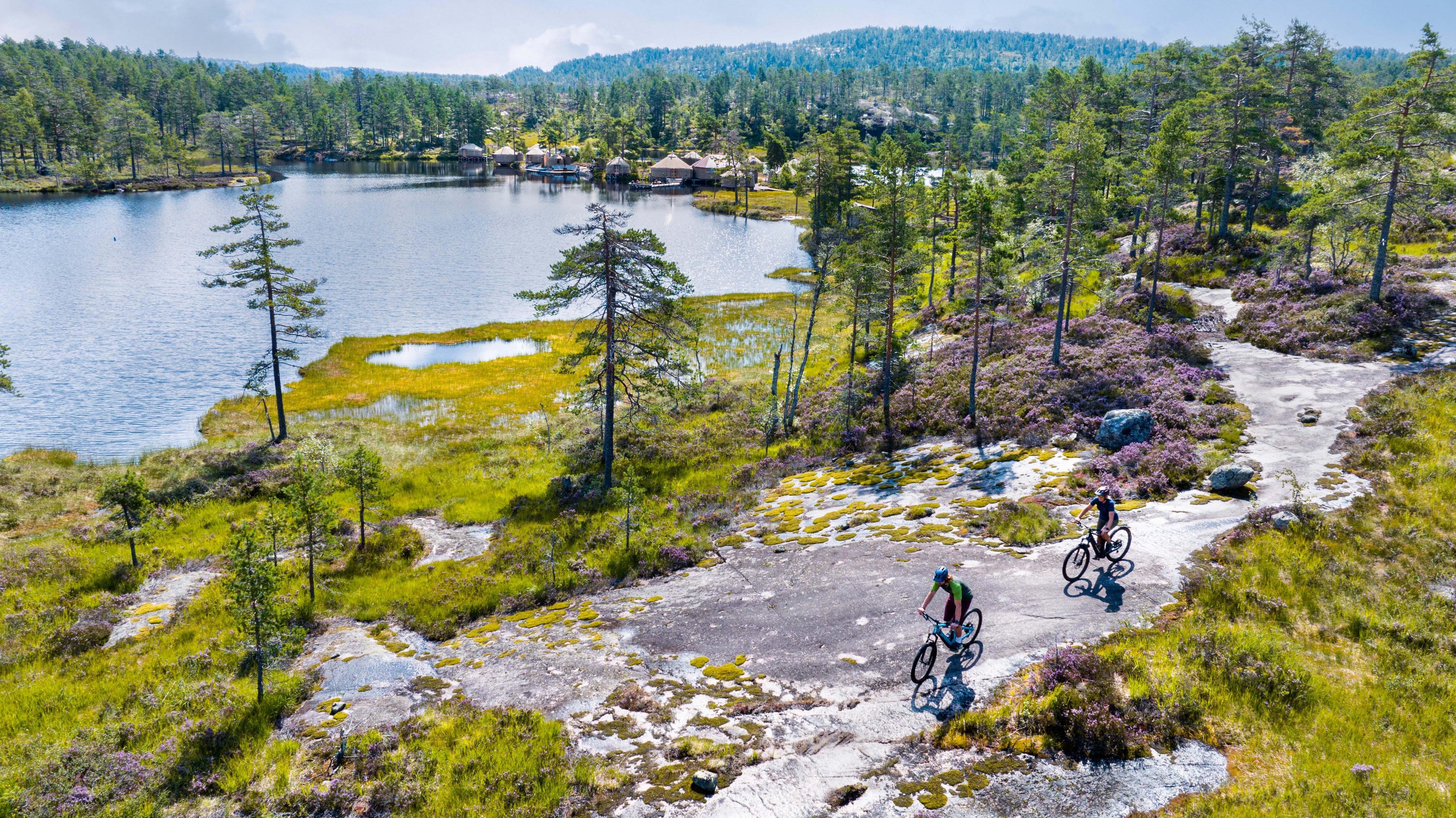 Two people riding Norrøna bikes on a path through a rocky, grassy area near a lake and forest. 
