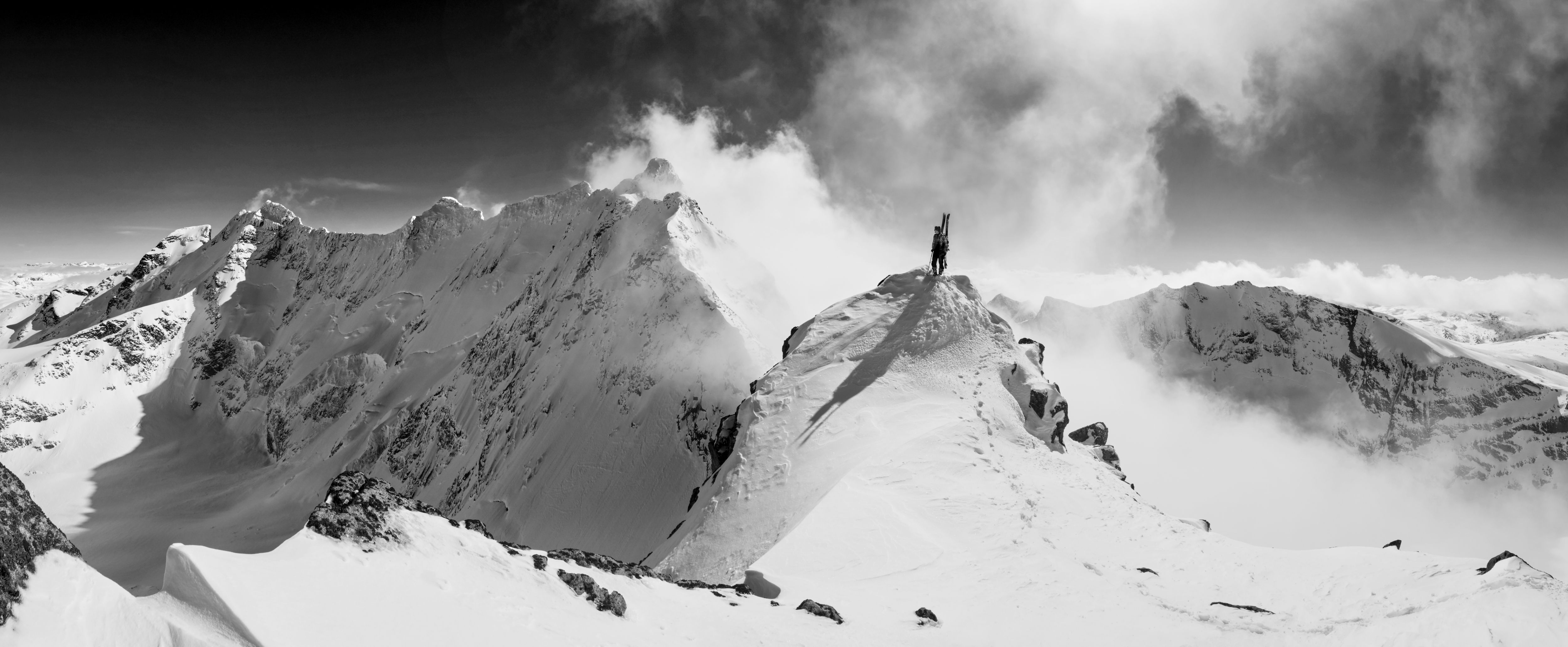 Black and white photo of a skier standing on a snowy mountain peak.  The skier is wearing Norrøna gear.  The sky is cloudy. 
