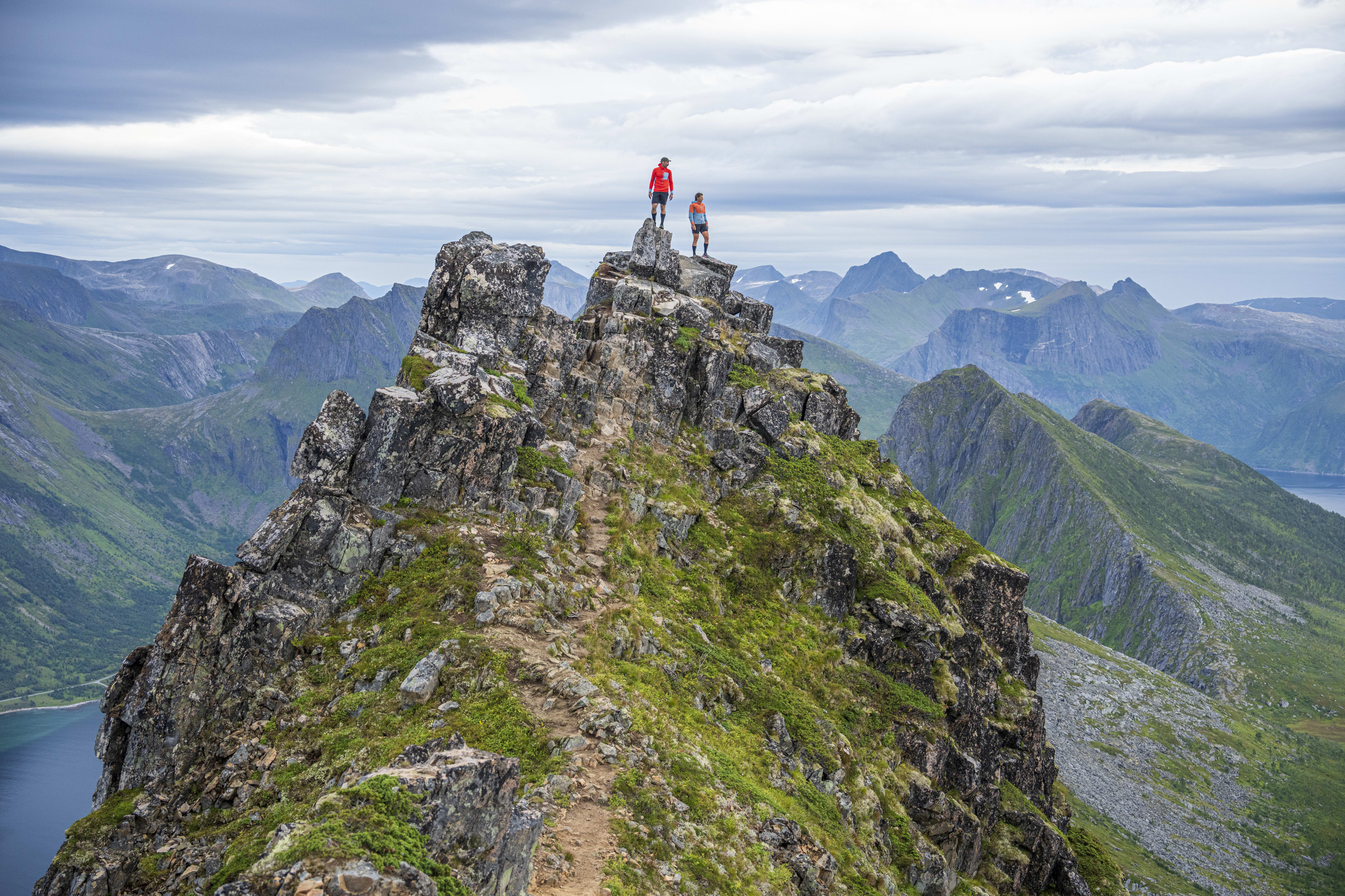 Two hikers stand on a rocky mountain peak overlooking a valley and fjord. The sky is cloudy. The hikers are wearing Norrøna clothing.