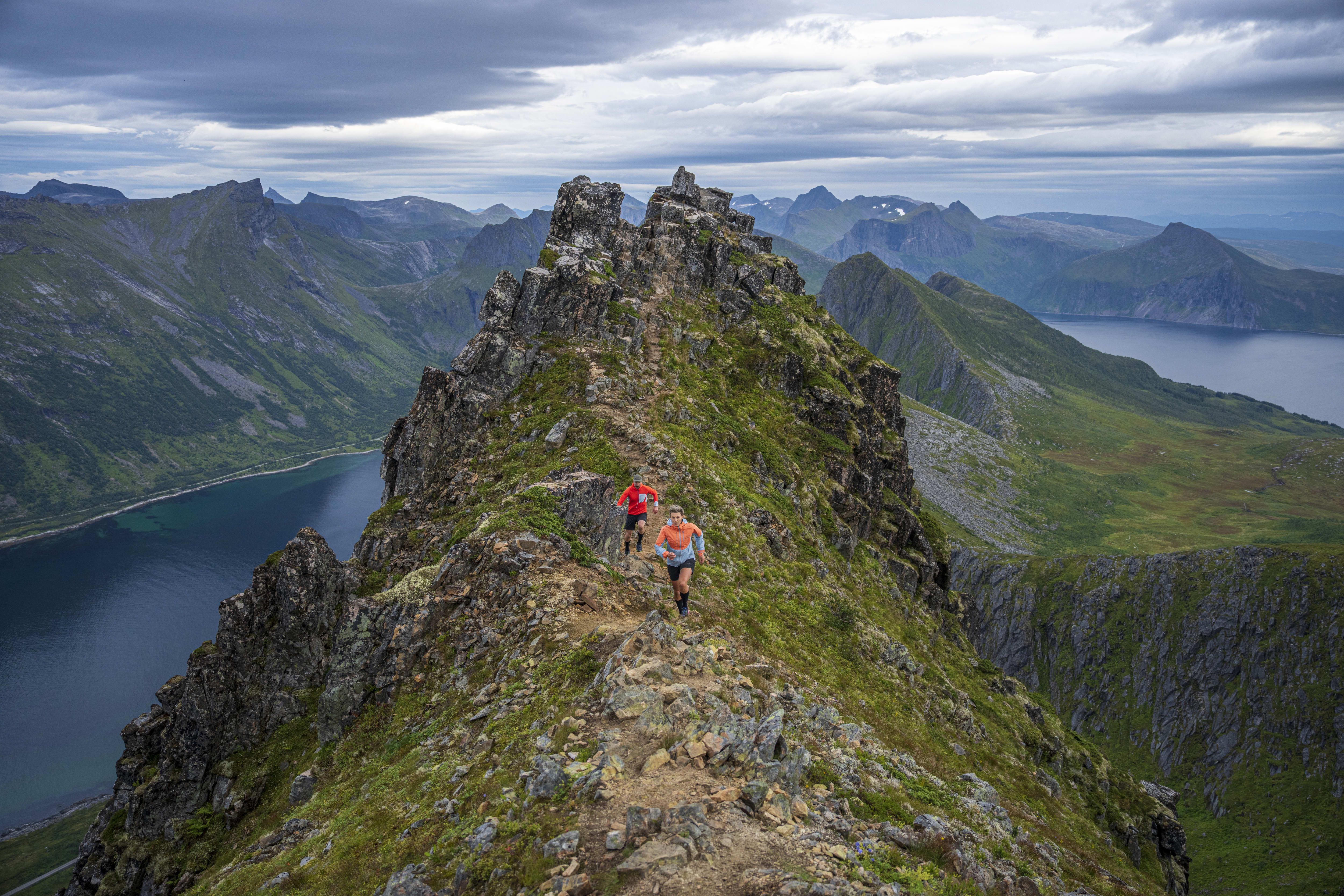 Two people wearing Norrøna gear hike along a narrow mountain path with a view of a fjord and a vast mountain range. 
