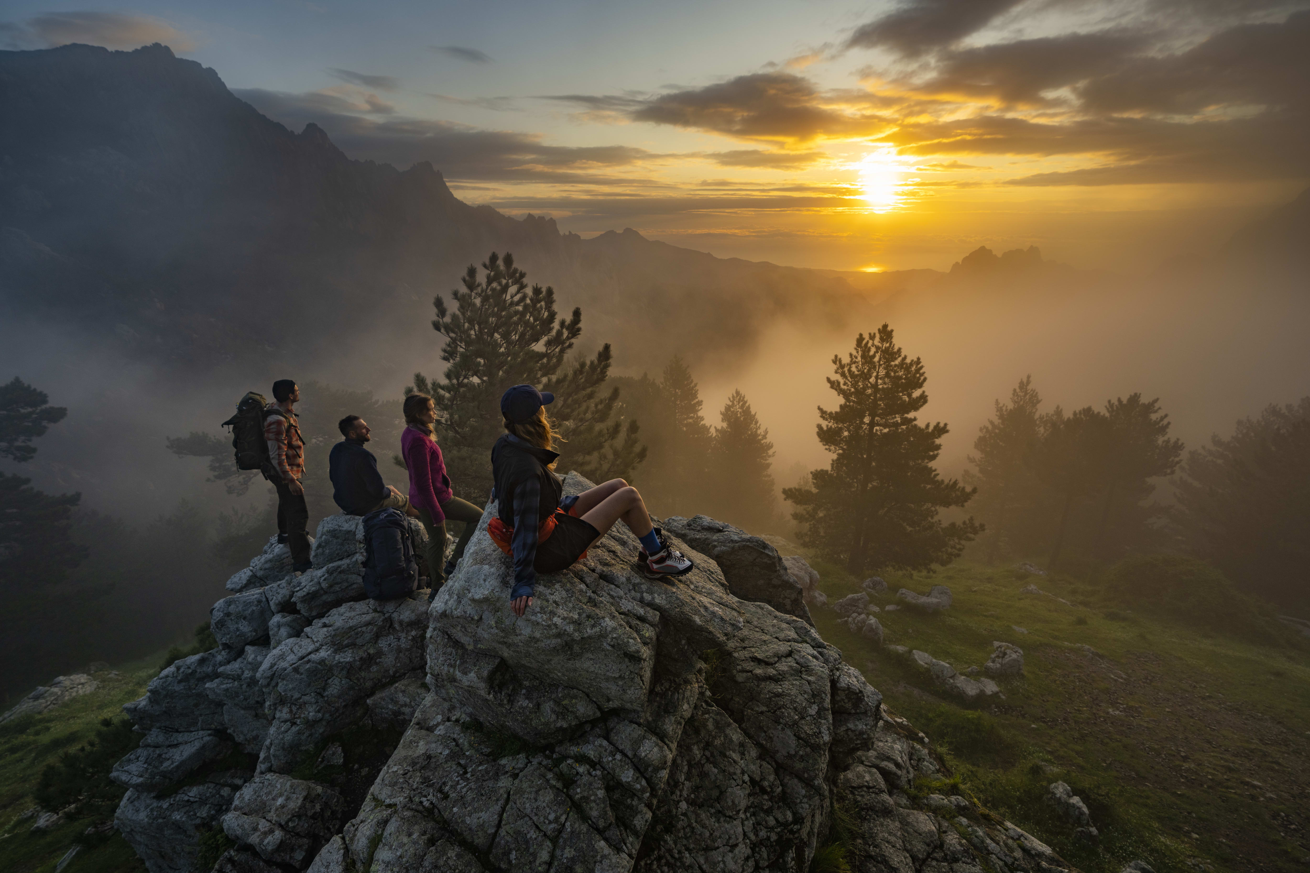 Four people stand on a rocky mountaintop, looking at a foggy sunset. The sun is a bright orange, behind a layer of fog that covers the valley below. There are trees in the foreground and background.