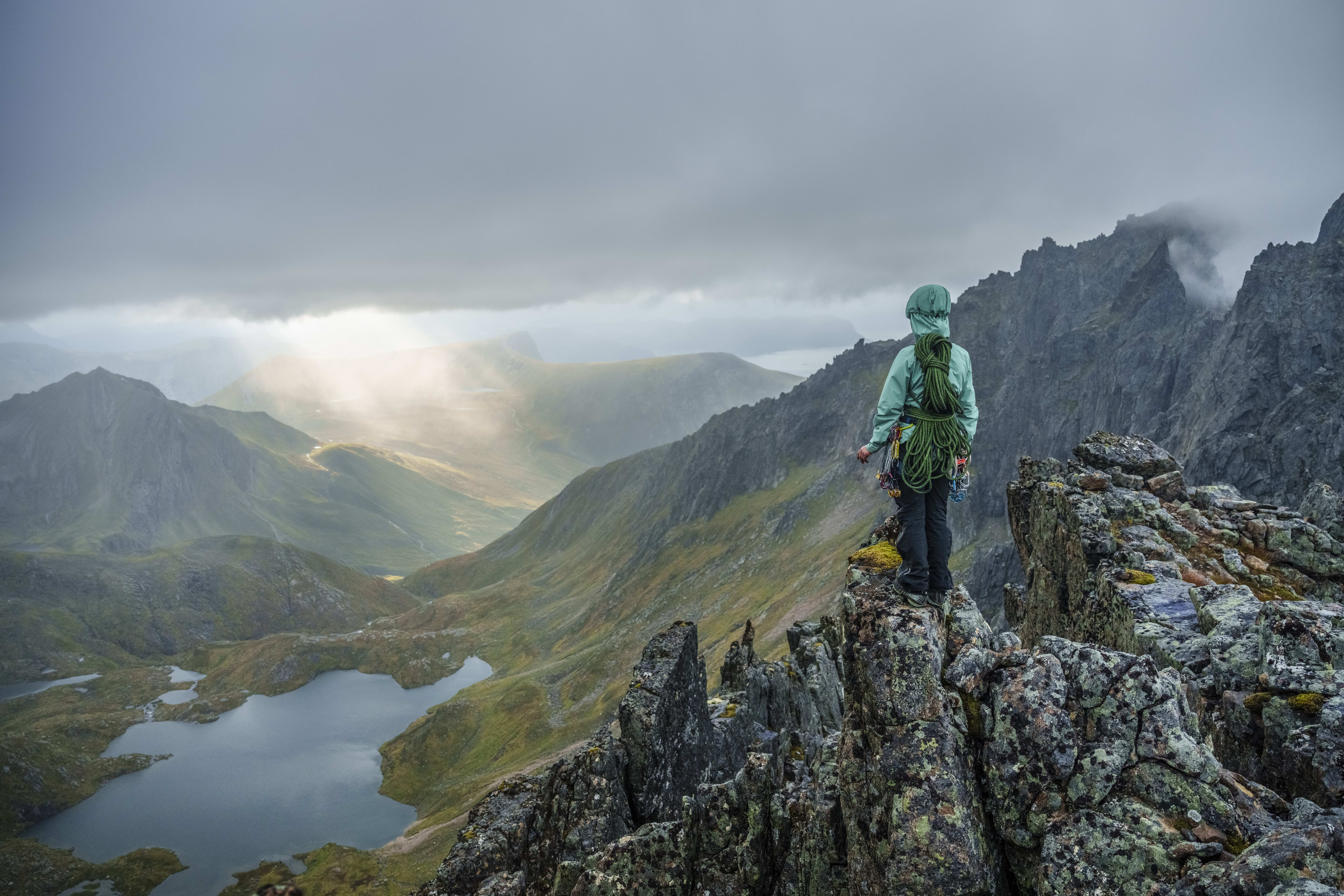 A hiker wearing Norrøna gear stands on a rocky mountain peak overlooking a valley with a lake. The sky is cloudy with rays of light shining through.