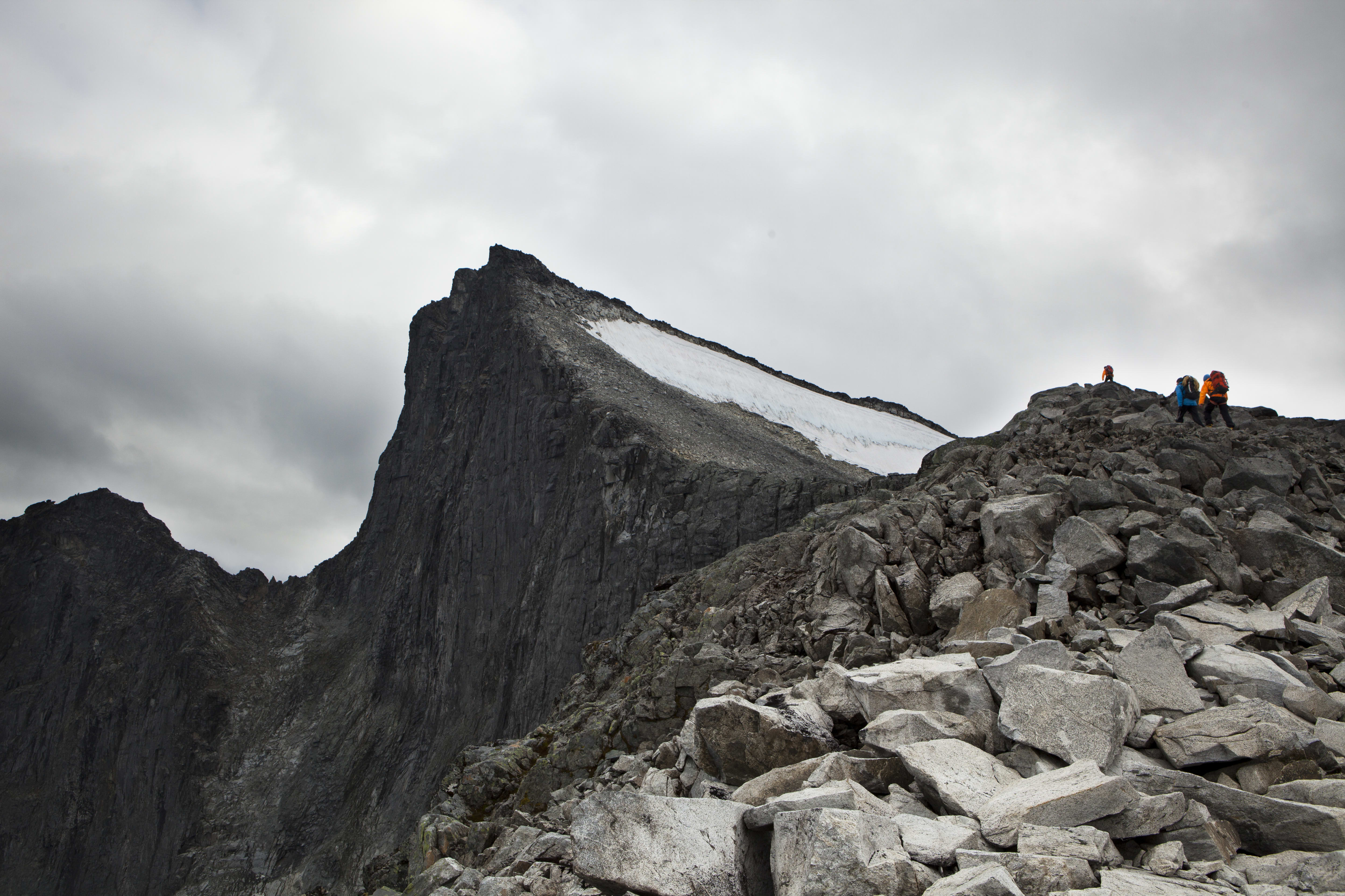 Three hikers on a rocky mountaintop with a glacier. The sky is cloudy. 

