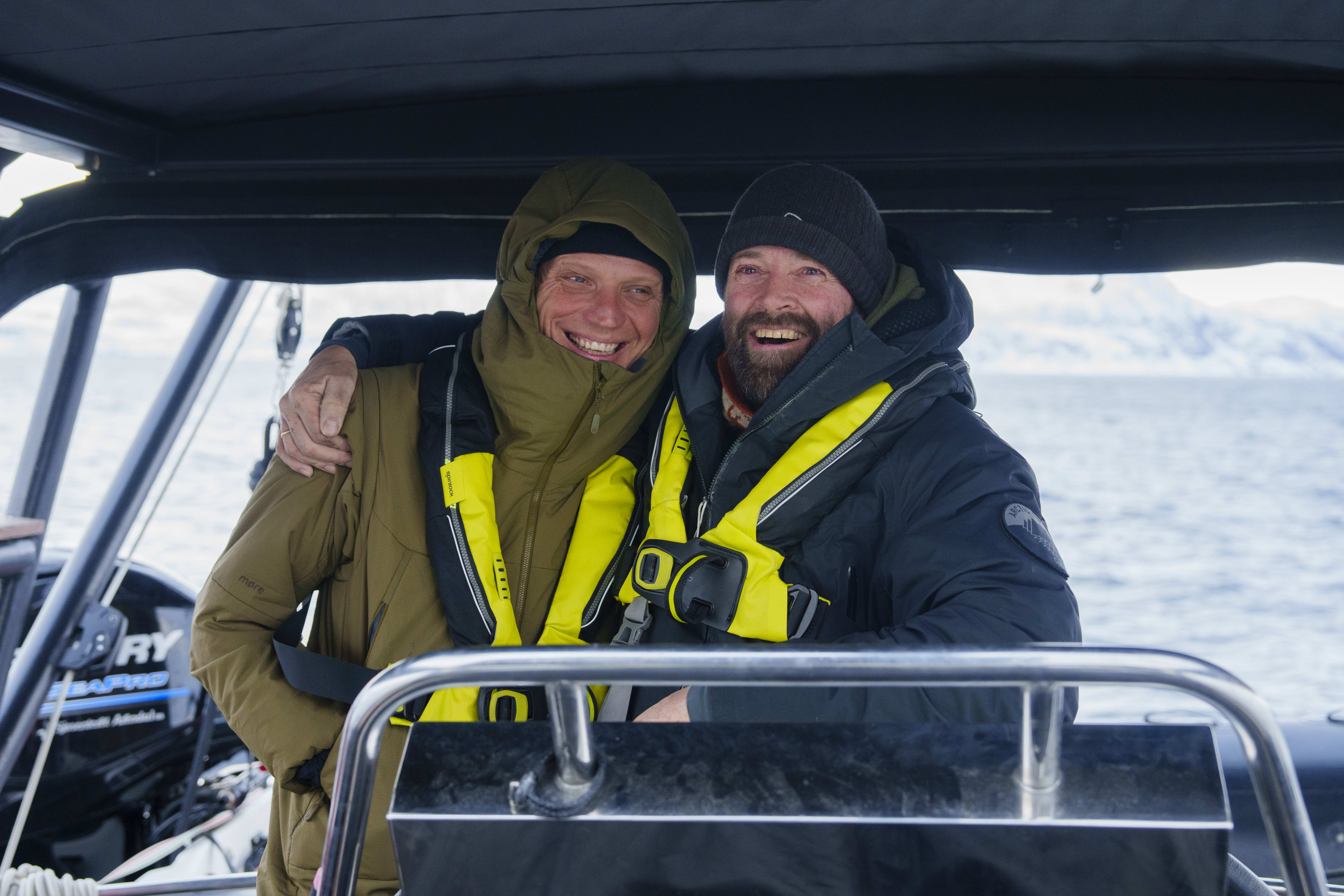 Two smiling men on a boat wearing Norrøna jackets and life vests. Water and mountains are in the background.