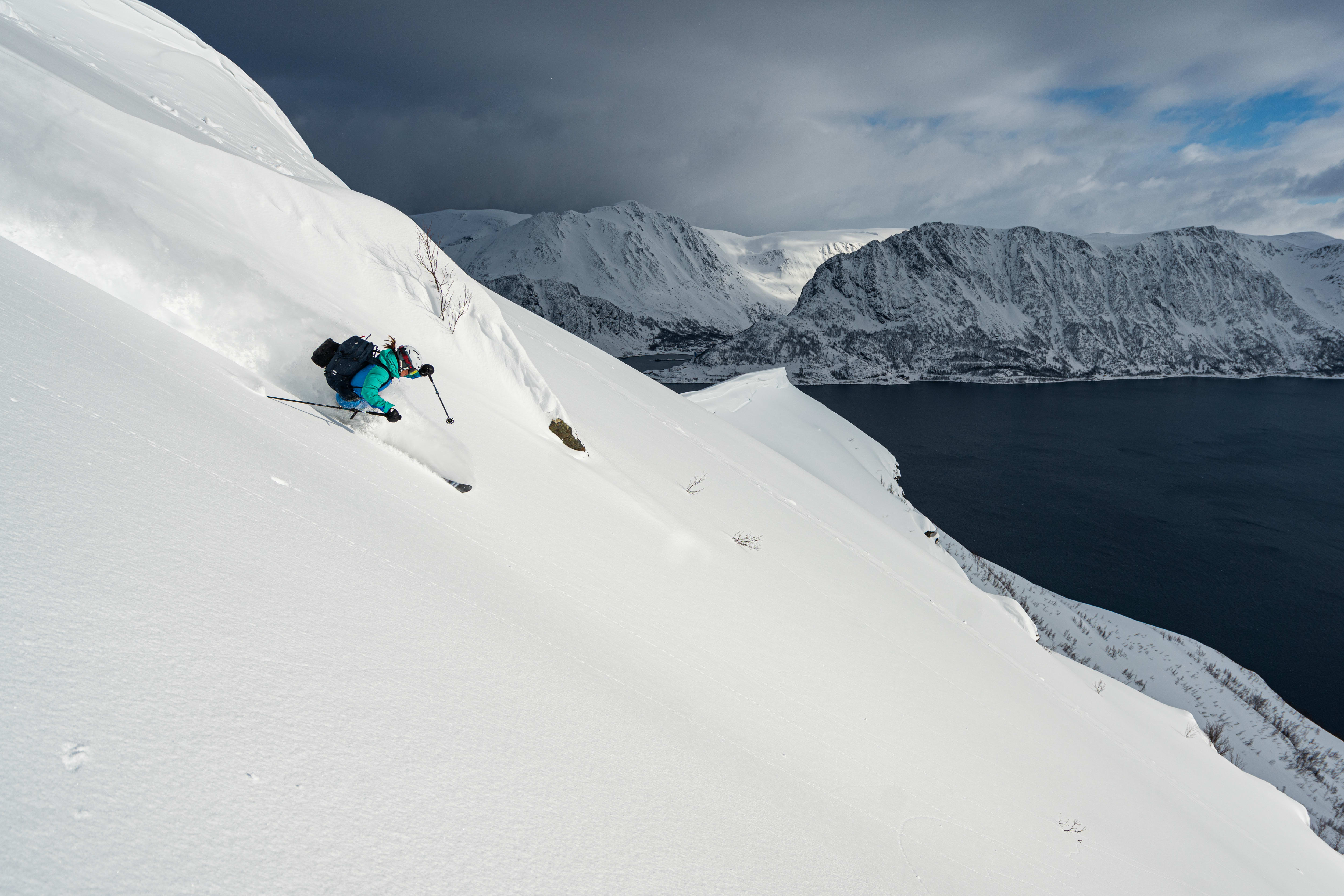 A skier in Norrøna gear makes a turn on a snowy mountainside overlooking a fjord. 
