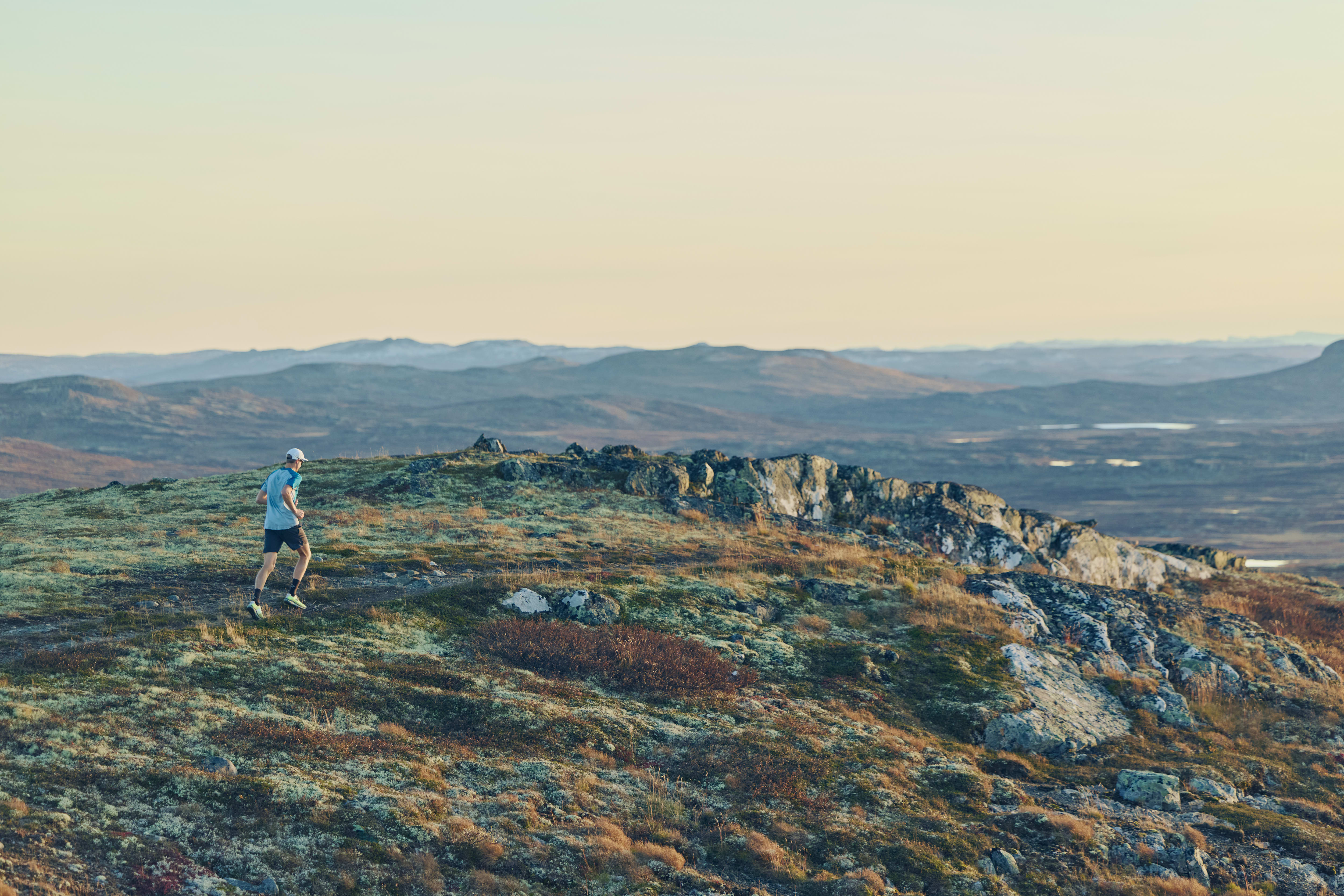 A person wearing a Norrøna shirt runs on a grassy, rocky mountaintop trail. Hilly landscape and sky are in the background.