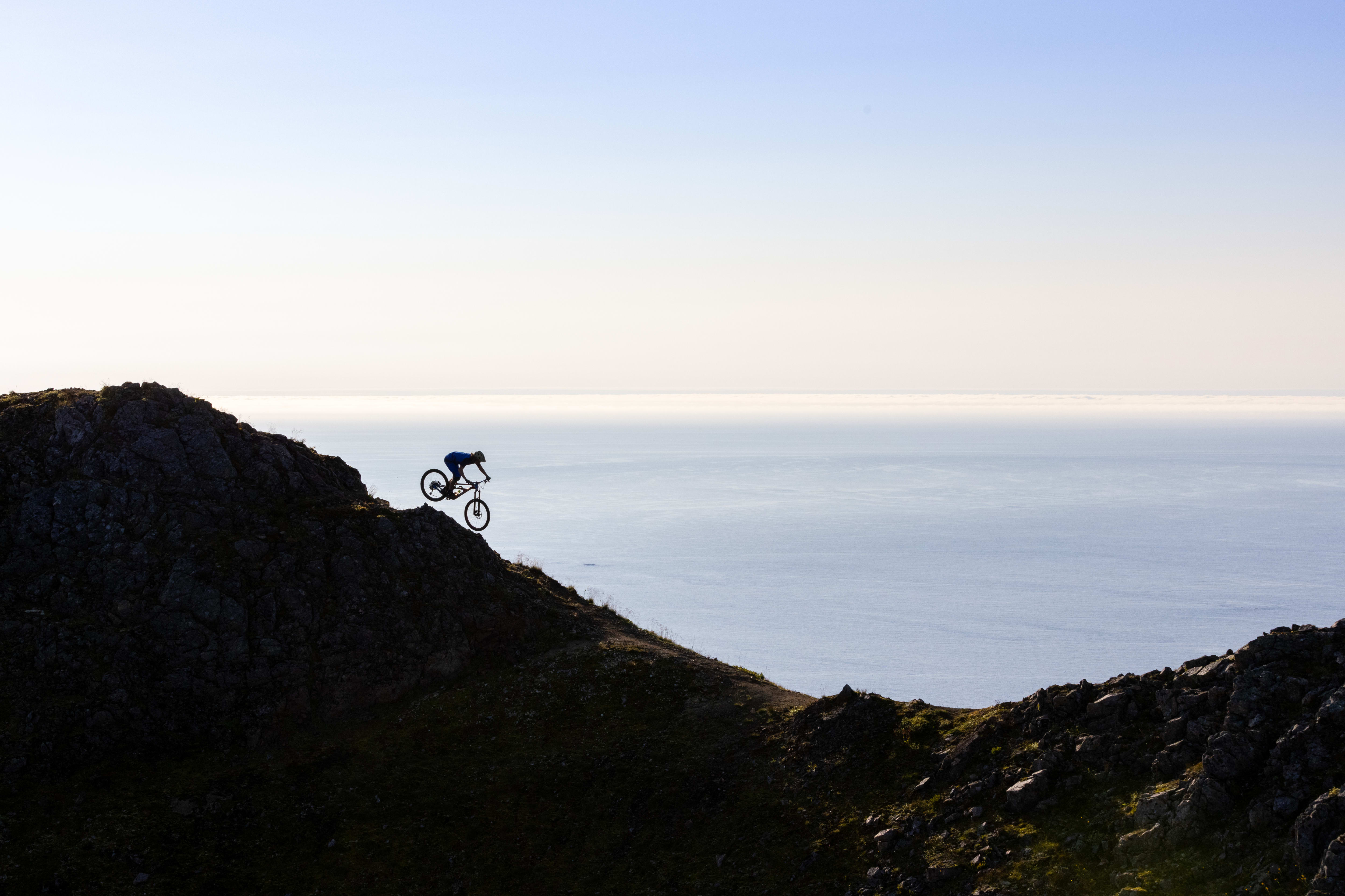 Silhouette of a person riding a Norrøna mountain bike down a rocky cliff overlooking the ocean.