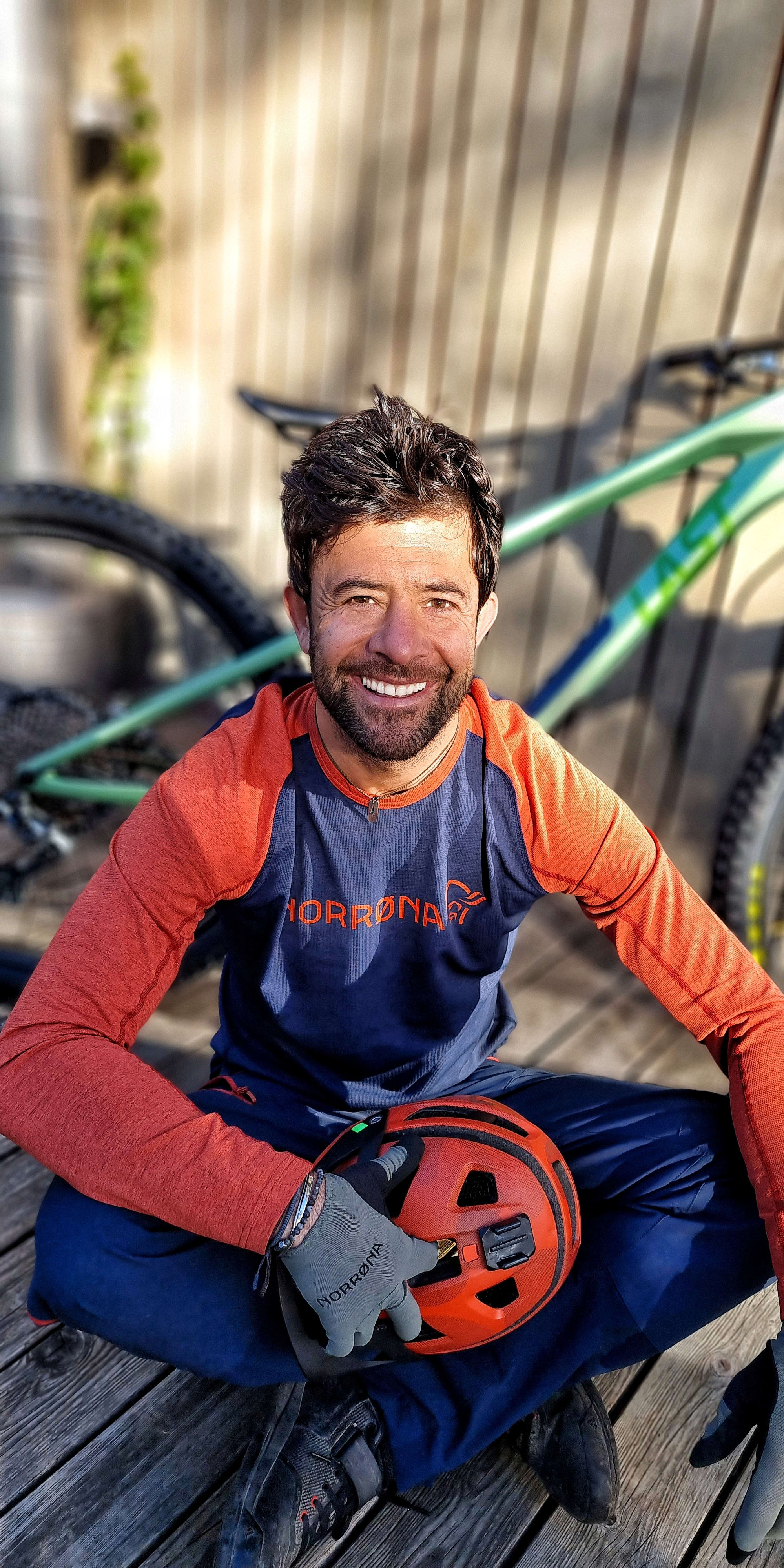 A man wearing an orange and blue long-sleeved shirt with a mountain logo is sitting on a wooden deck with his hands resting on his legs. He is holding a red bicycle helmet with a camera mount. A mountain bike is blurred in the background.