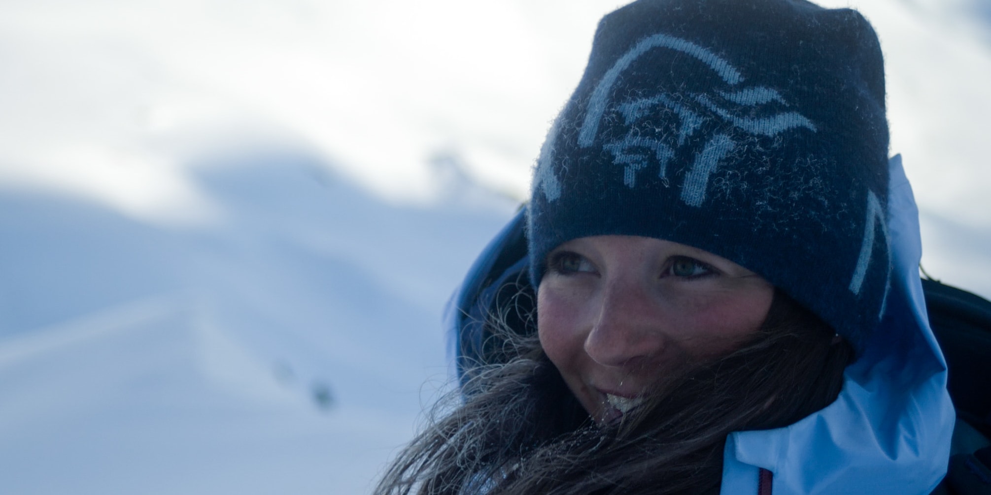 A smiling woman in a Norrøna hat and blue jacket looks left against a snowy mountain backdrop.