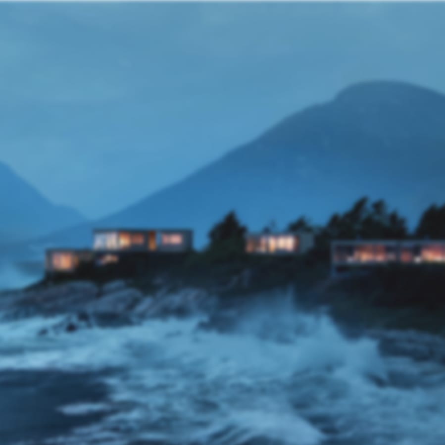 Illuminated modern cabins on a rocky coast with crashing waves and mountains at twilight.