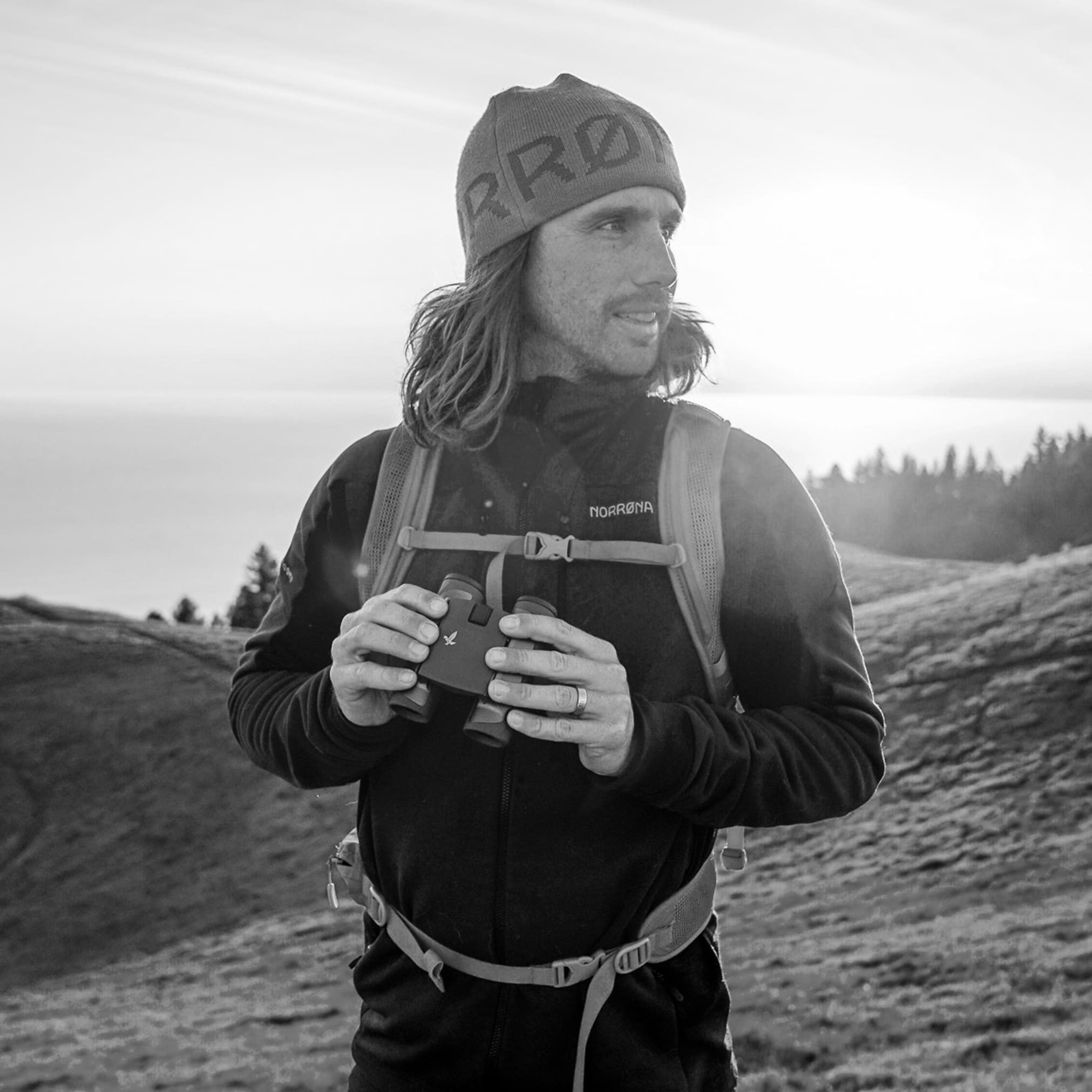 A man wearing a Norrøna jacket and a hat holds binoculars while looking to the side on a hill. 
