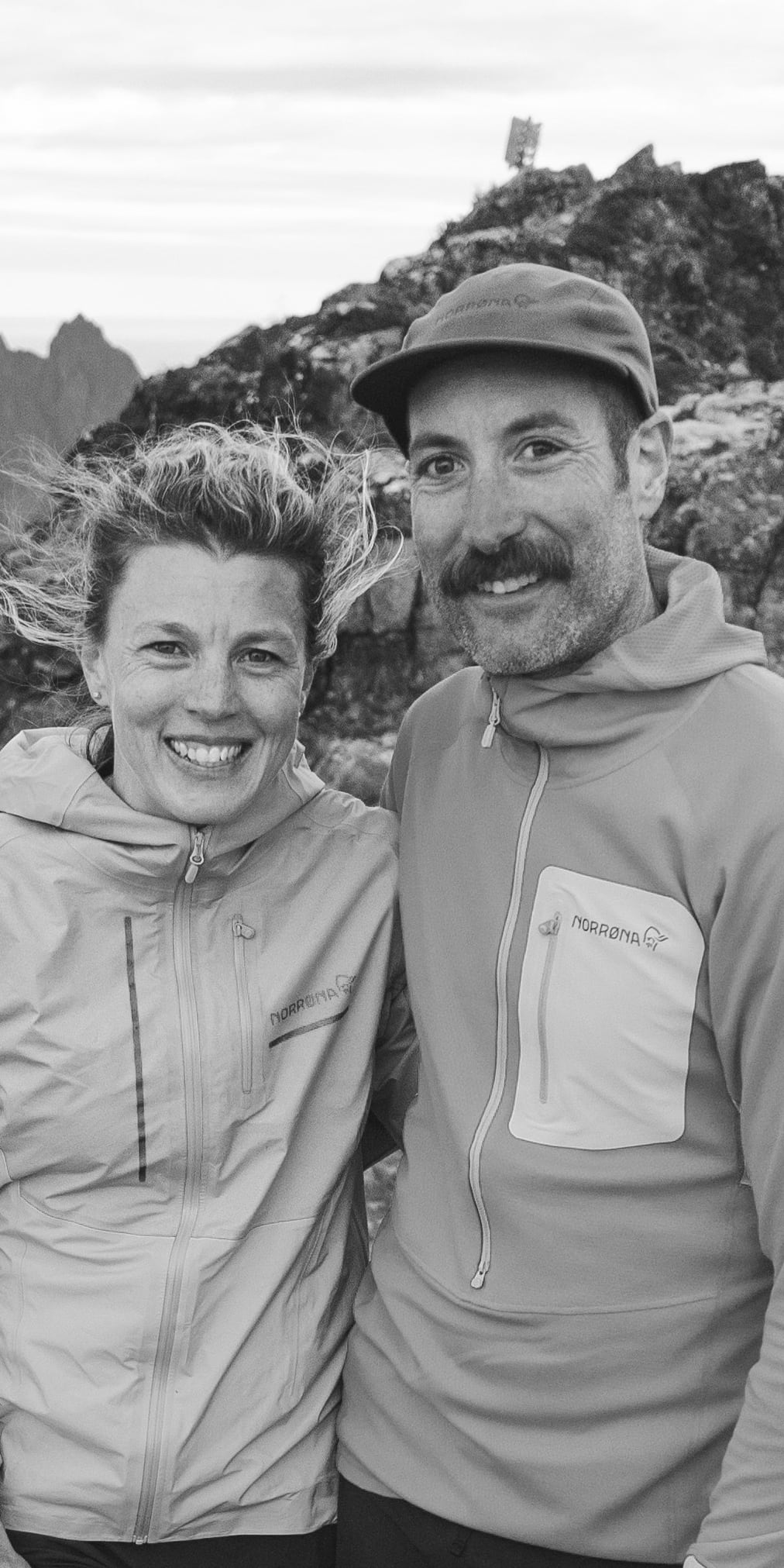 Two people wearing Norrøna jackets stand on a rocky mountaintop, smiling, overlooking a body of water. 
