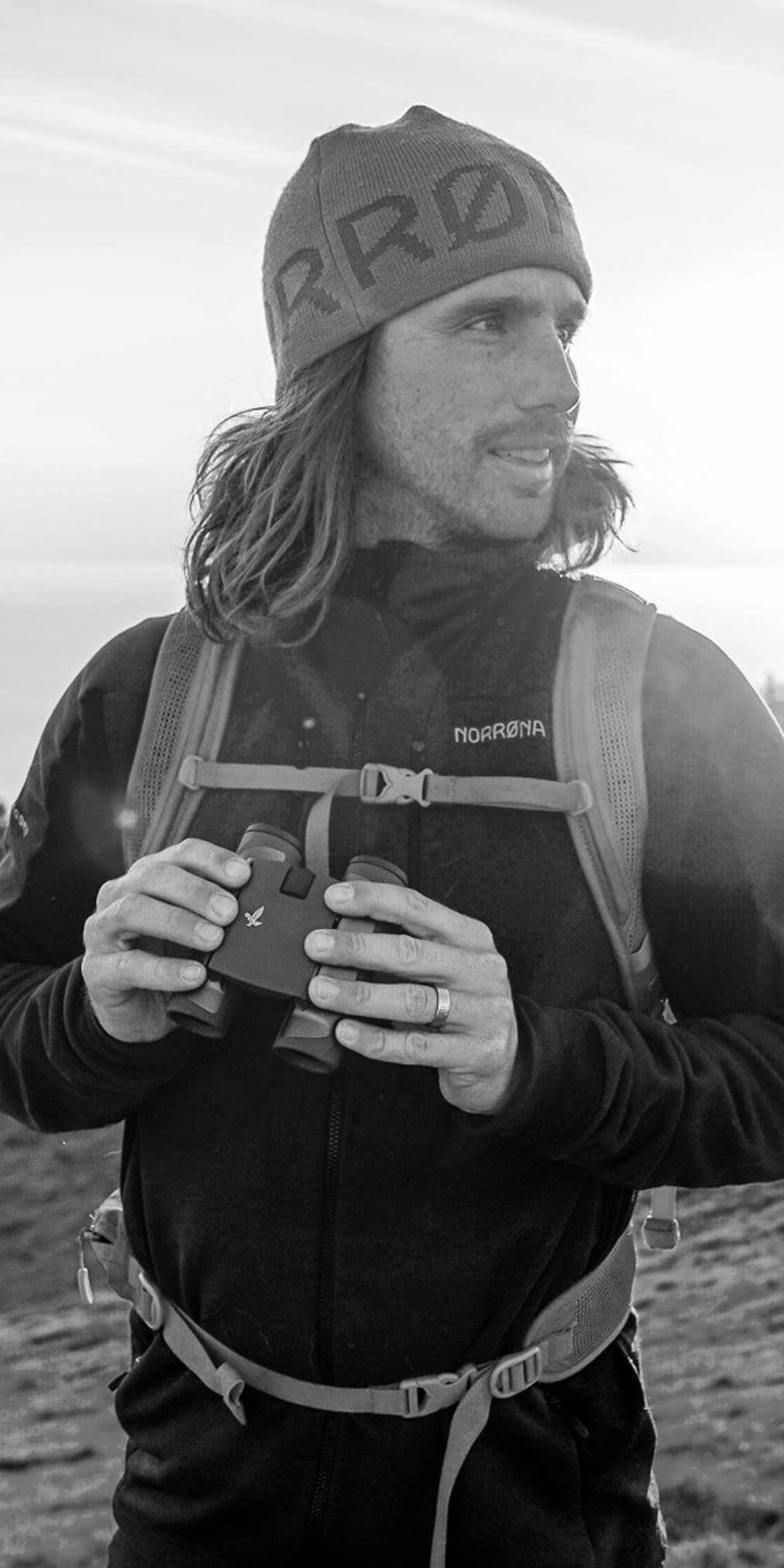 A man wearing a Norrøna jacket and a hat holds binoculars while looking to the side on a hill. 
