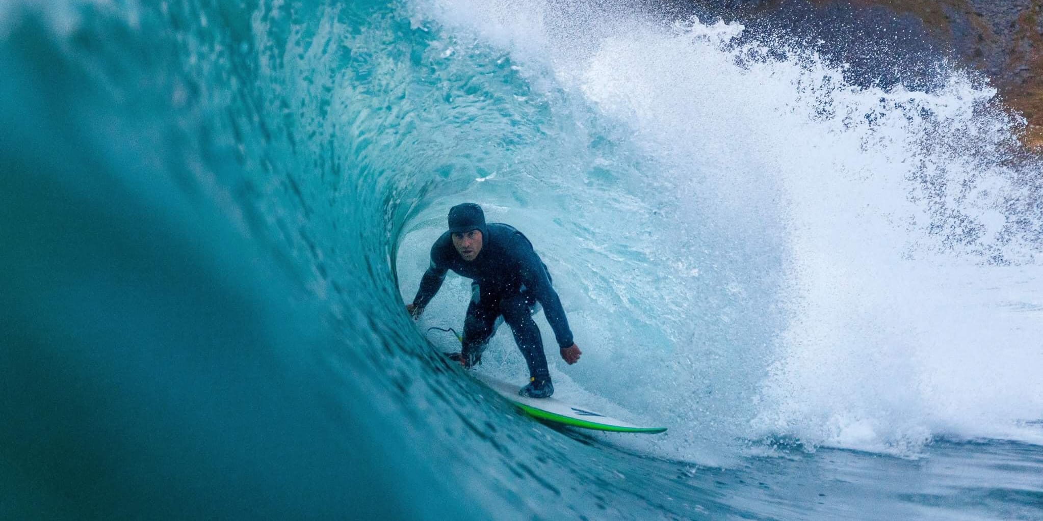 A surfer in a black Norrøna wetsuit rides inside a turquoise wave, with white spray and a rocky coastline visible in the background.