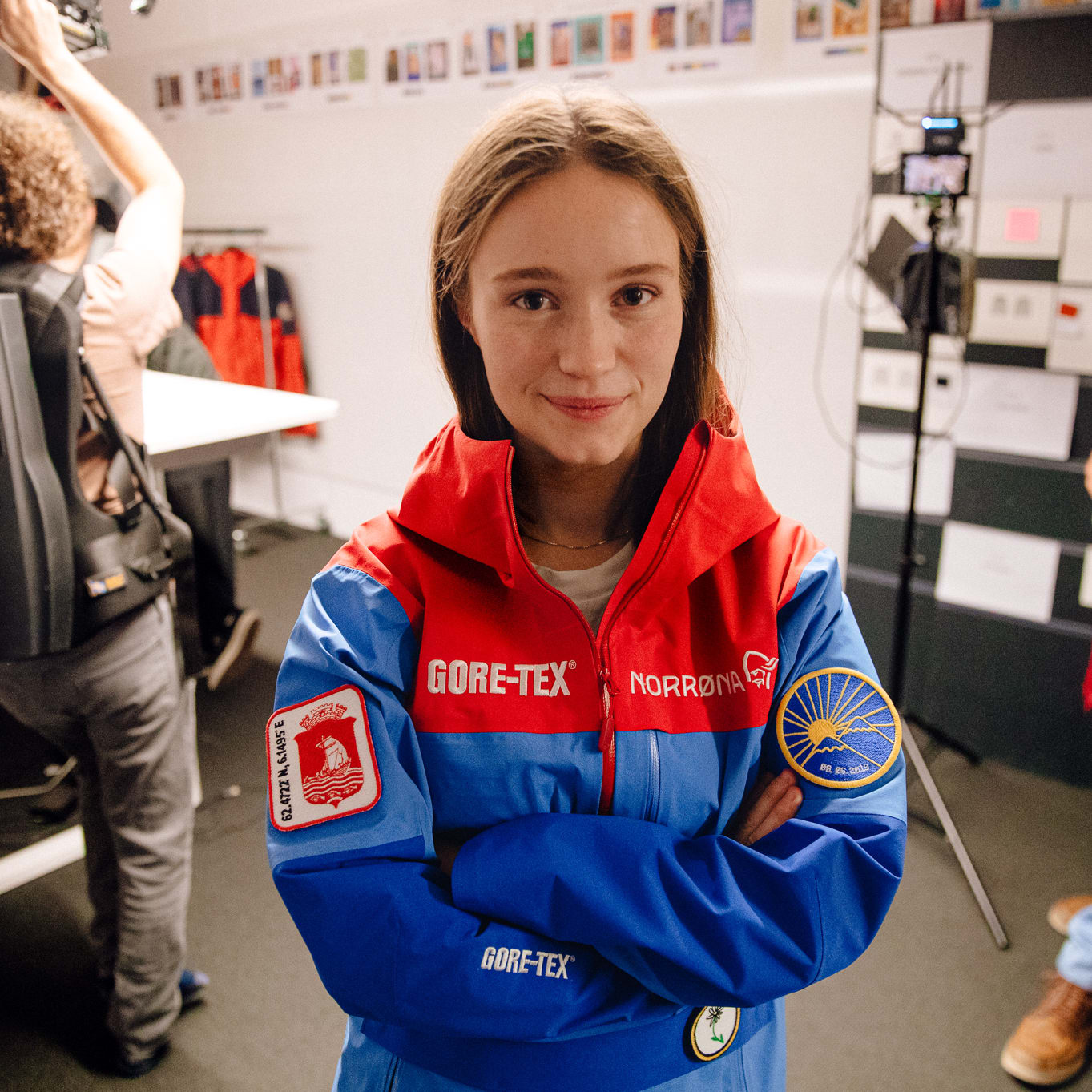 A woman wearing a blue Norrøna jacket with a red hood, arms crossed, smiling. She has a red and blue patch on her left sleeve and a yellow patch with a sun on her right sleeve. 
