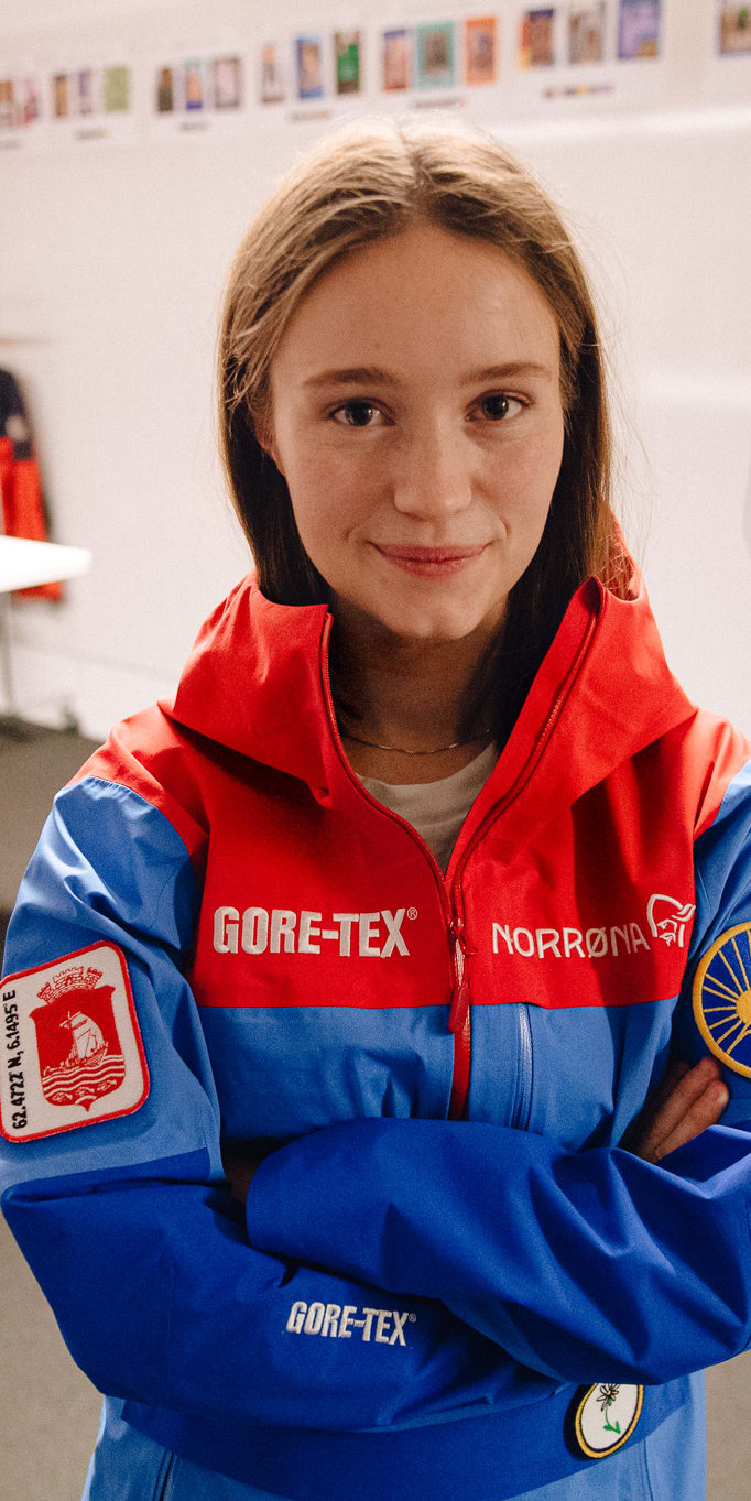 A woman wearing a blue Norrøna jacket with a red hood, arms crossed, smiling. She has a red and blue patch on her left sleeve and a yellow patch with a sun on her right sleeve. 

