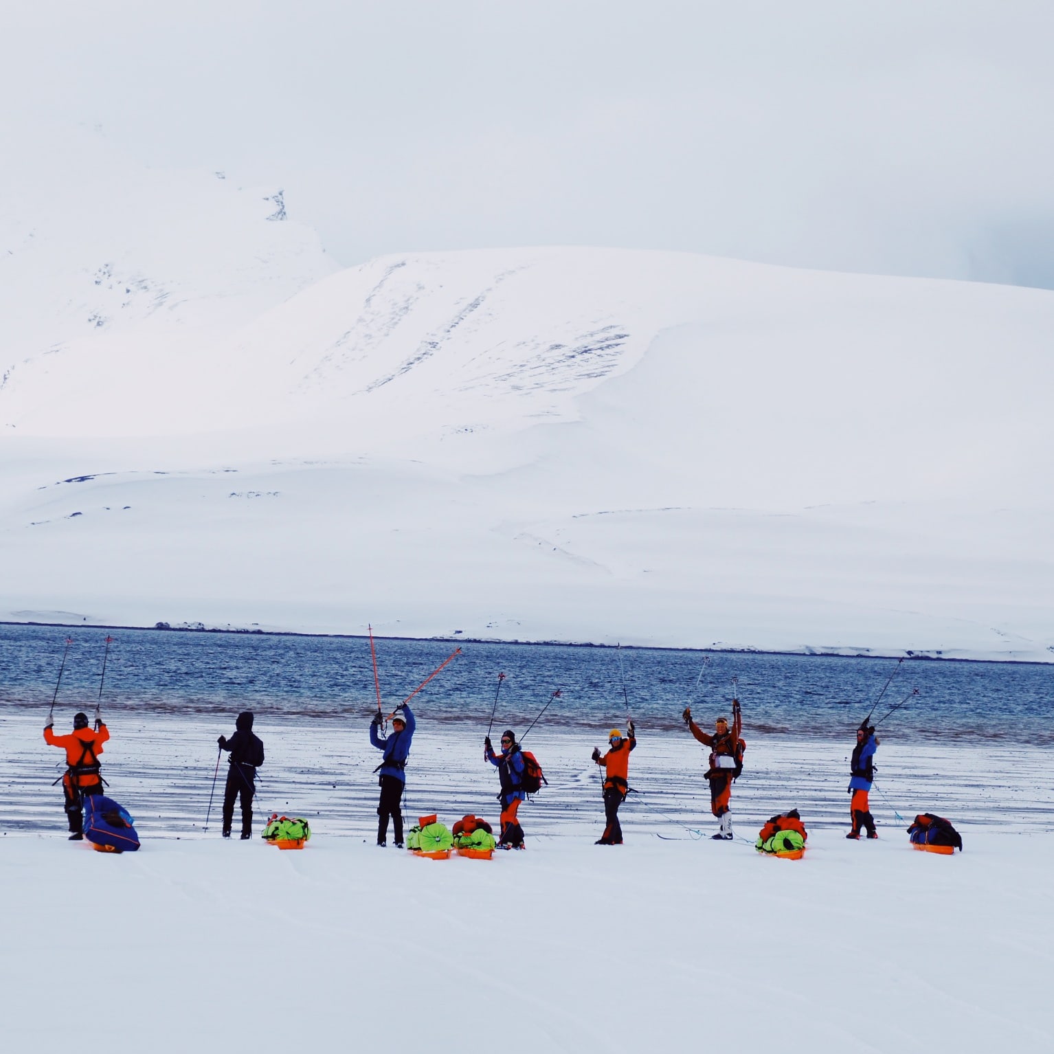 A group of eight people in winter gear, stand on a snowy beach in front of a snow-covered mountain range. They are holding poles and some have backpacks on.
