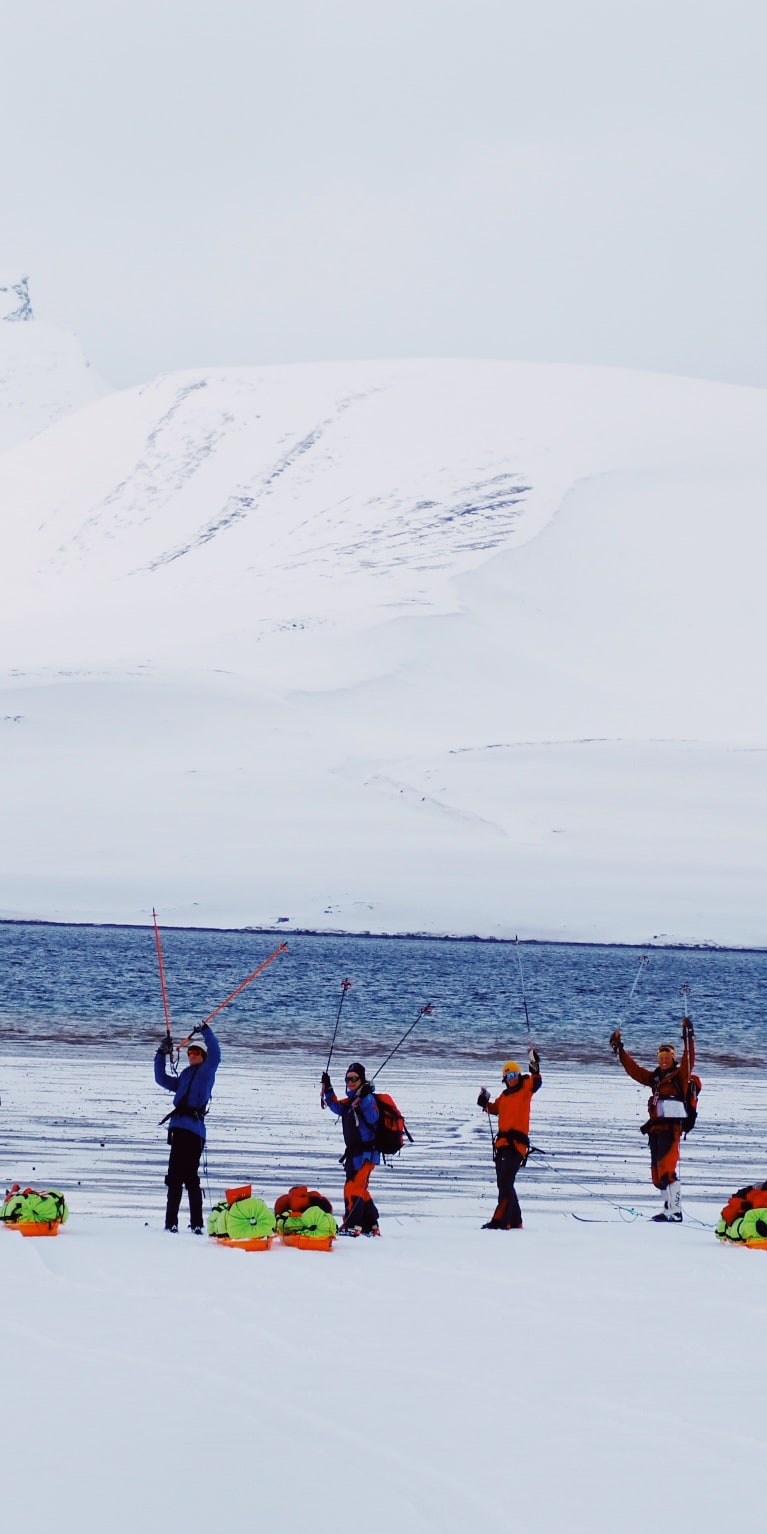 A group of eight people in winter gear, stand on a snowy beach in front of a snow-covered mountain range. They are holding poles and some have backpacks on. 
