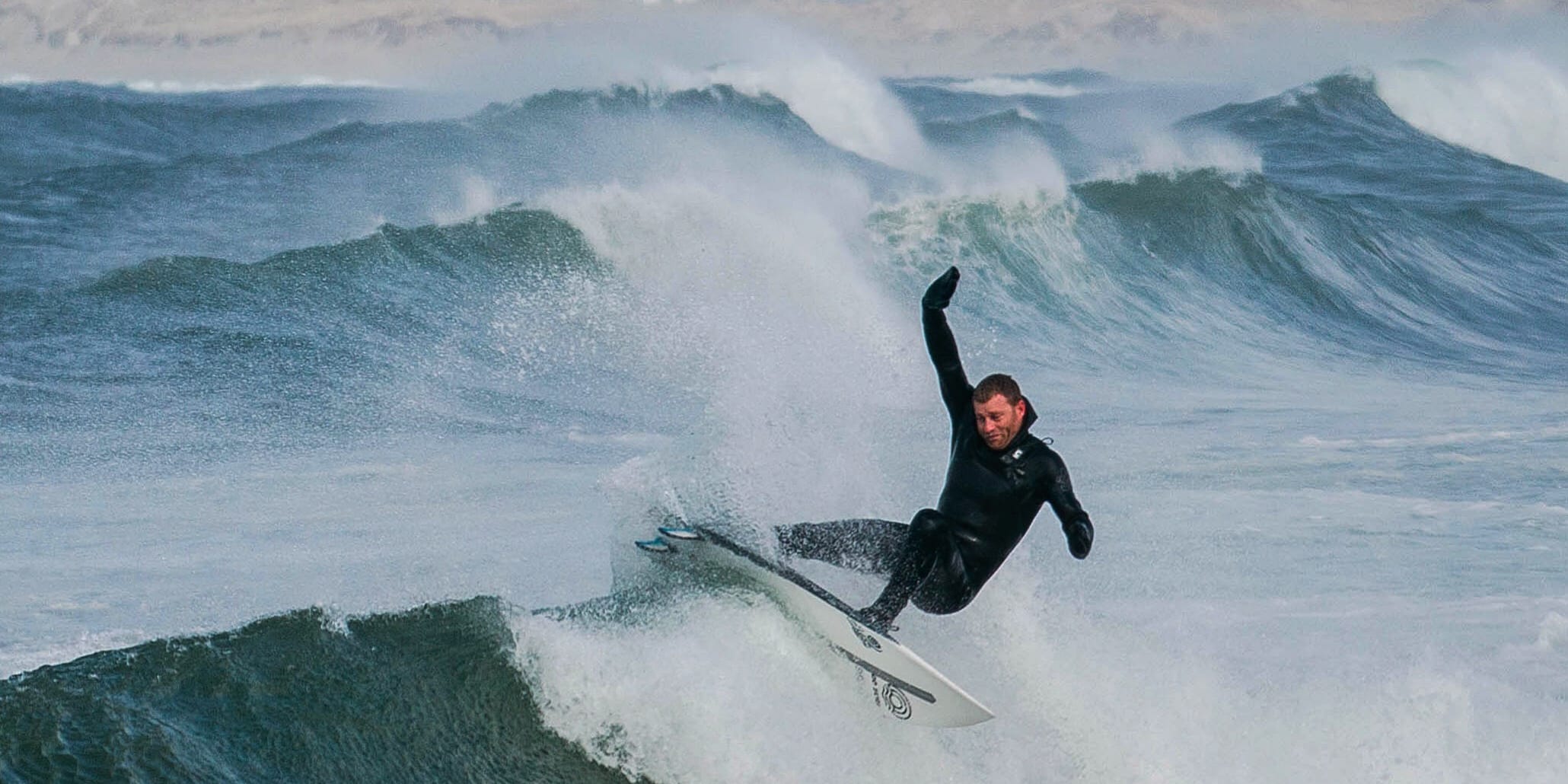 A surfer in a Norrøna wetsuit rides a large wave with white spray against an overcast sky. They are angled upwards, one arm raised.
