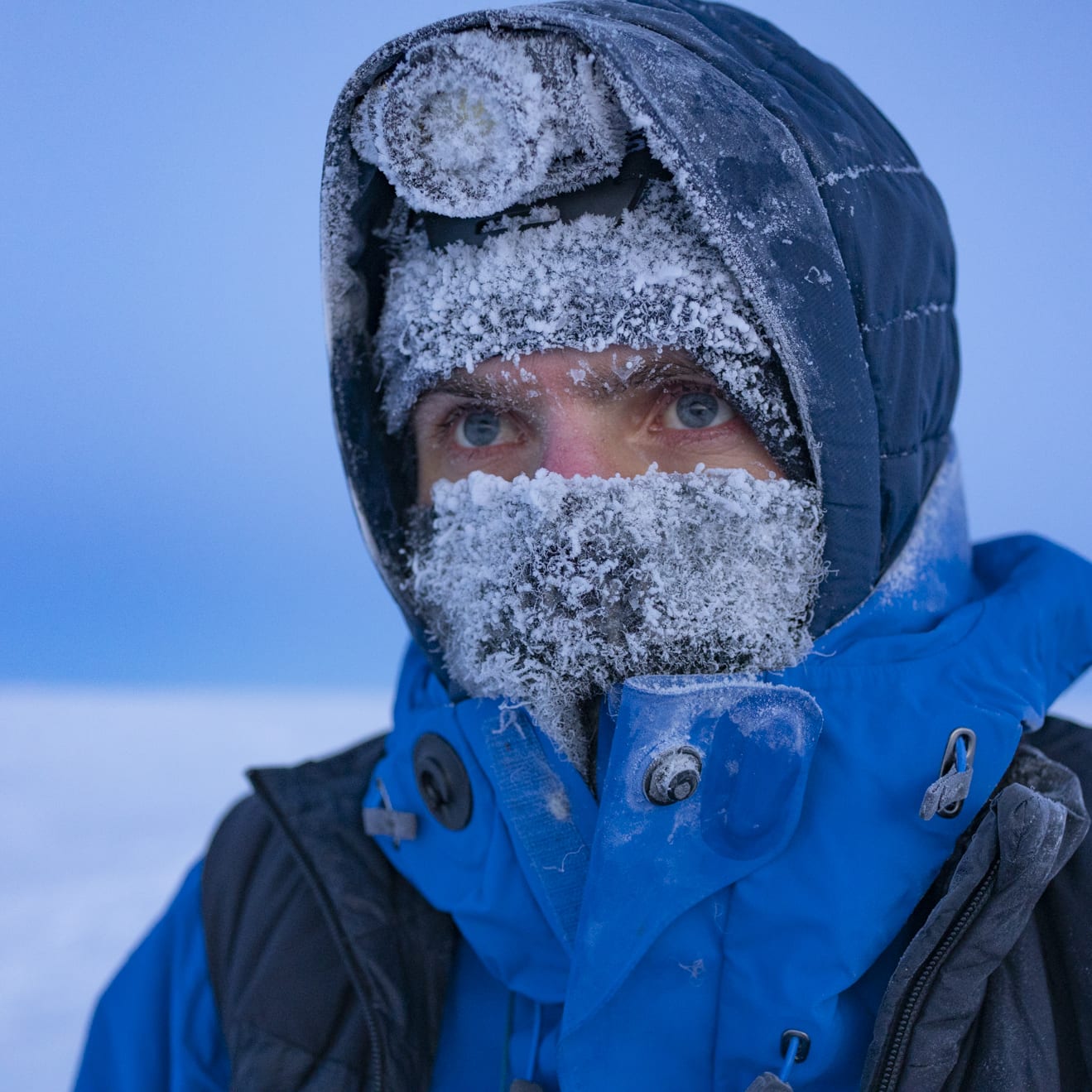 A person wearing a blue Norrøna jacket, hood, and a black Norrøna vest, is covered in frost. Their face is obscured by the frost and their eyes are visible.