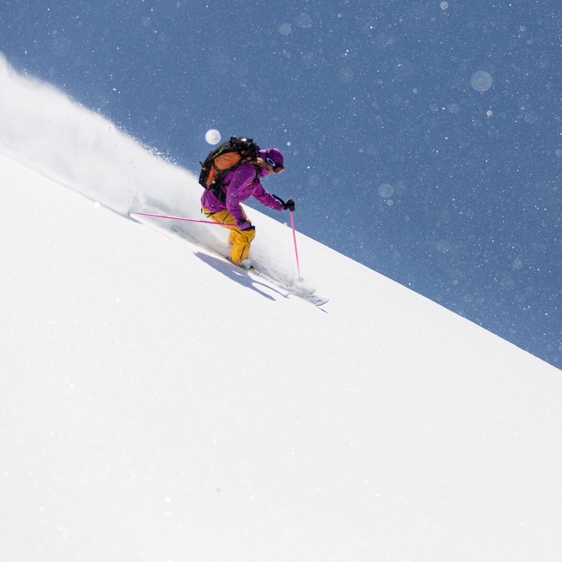 A skier in Norrøna purple jacket and yellow pants skis down a snow-covered slope against a blue sky.
