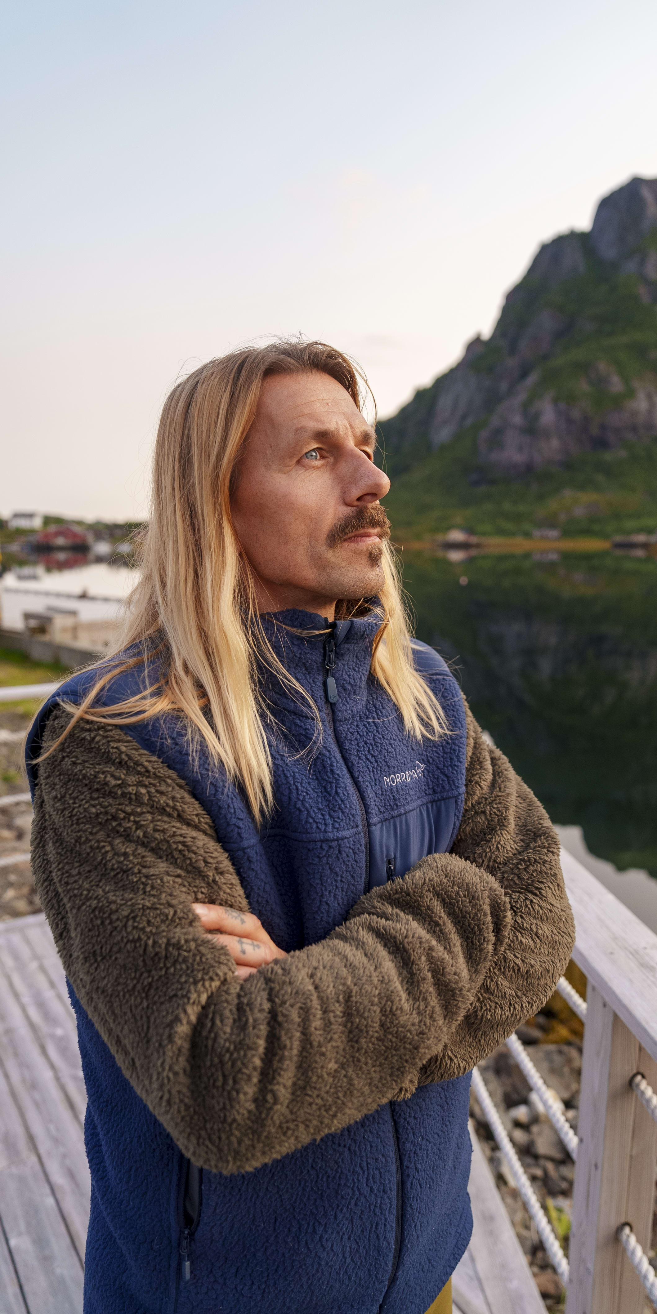 Man with long blonde hair wearing a blue Norrøna fleece jacket, standing on a wooden pier, looking out over a body of water and mountains. 
