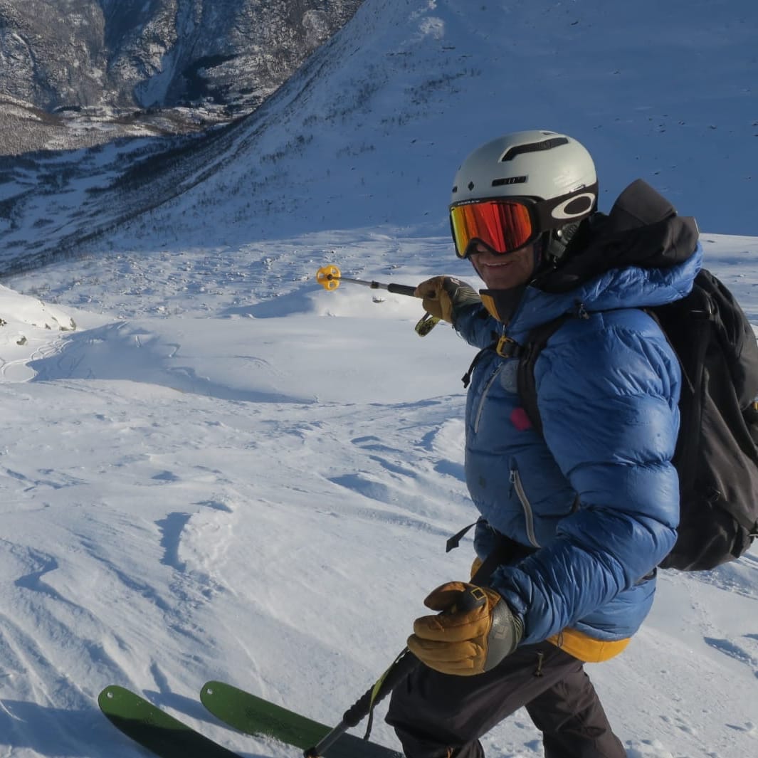 A person in a Norrøna jacket and ski gear stands on a snowy mountainside, holding a ski pole.  The view is looking down a snow-covered slope towards a mountain range in the distance. 

