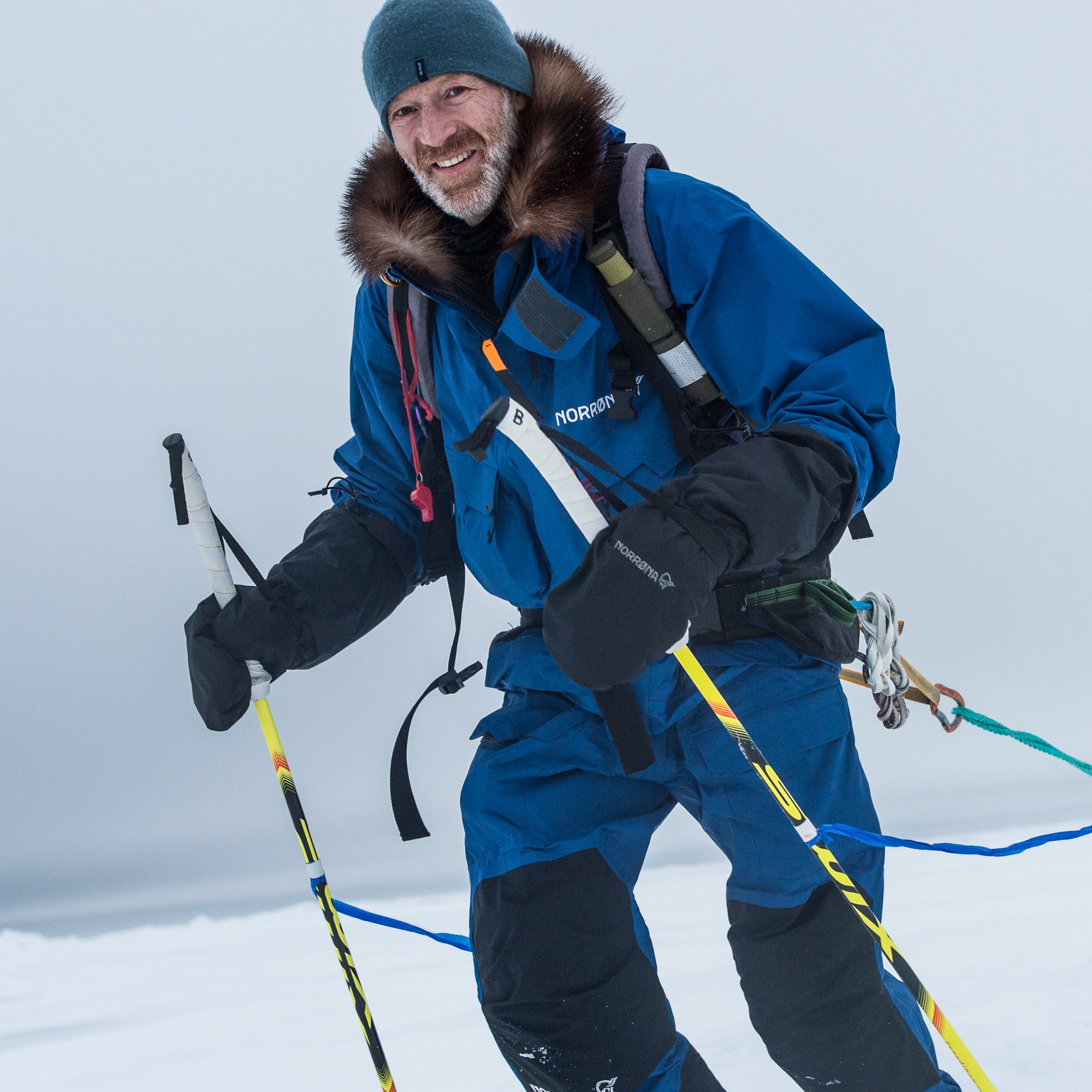 A man in a blue Norrøna jacket and black gloves skis on snow. He holds two ski poles and pulls a sled behind him.  
