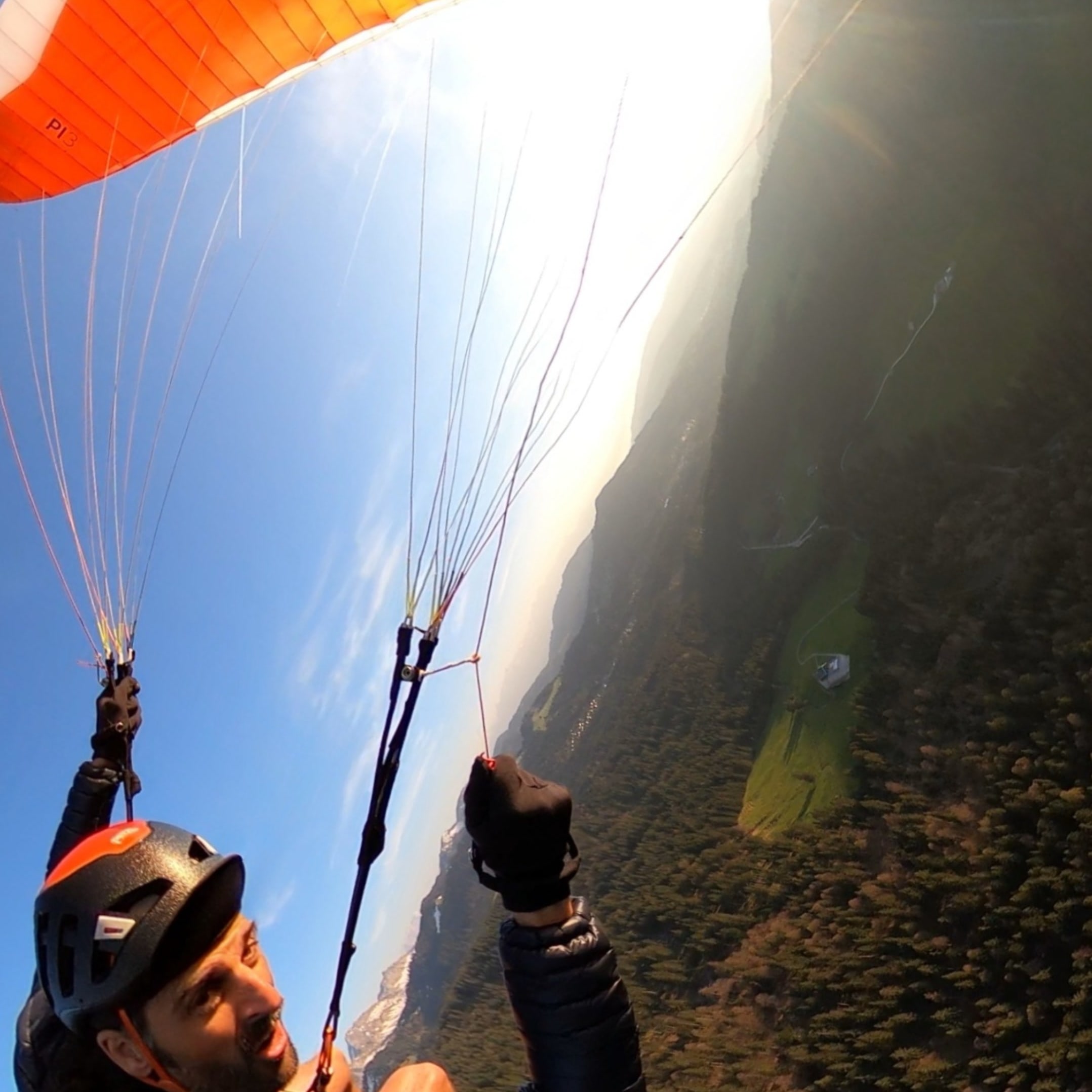 A person wearing a helmet and a black jacket is paragliding over a forest. The paraglider is orange and white with the number 13 on it. The sky is blue and the sun is shining.