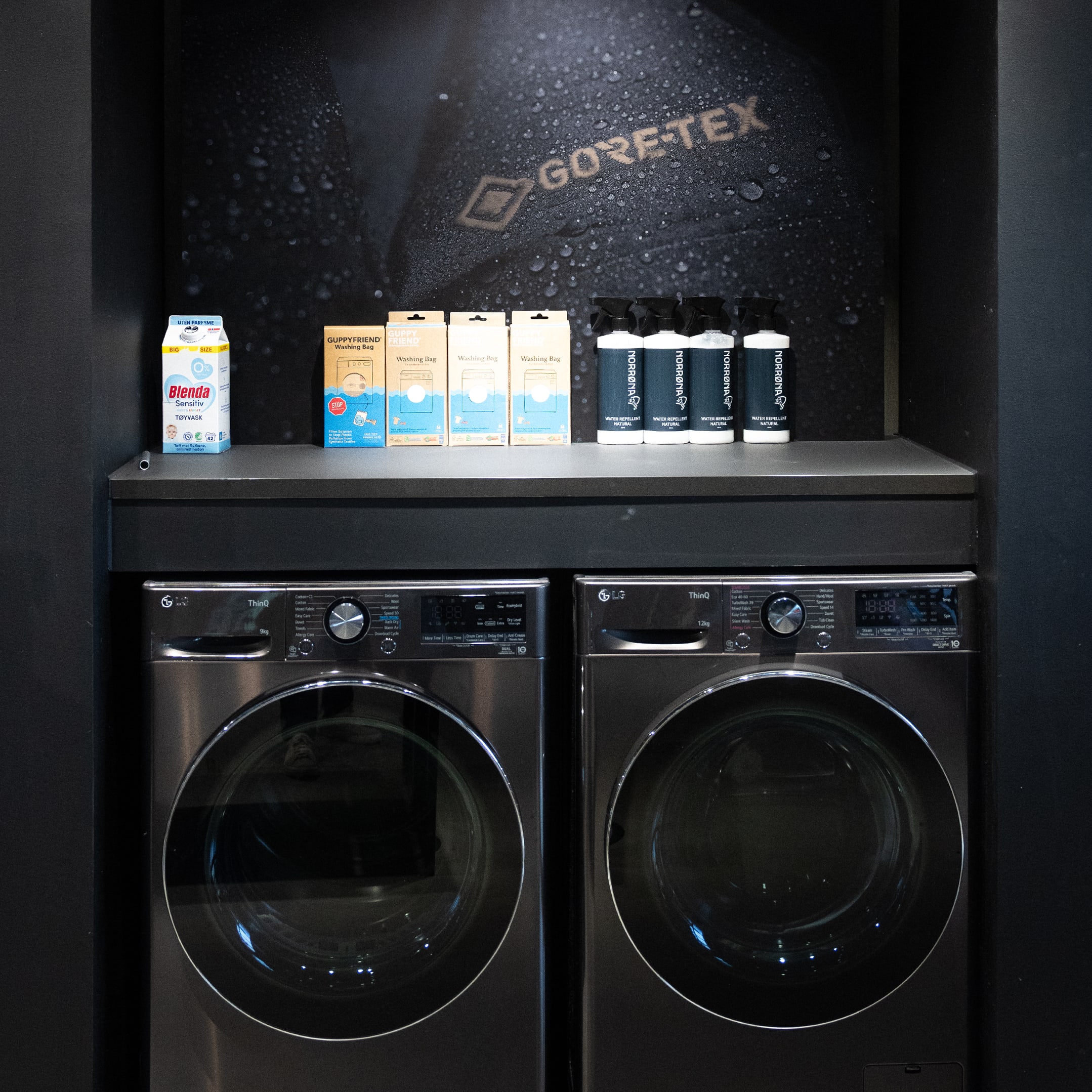 Two LG washing machines sit below a shelf displaying laundry products, including Norrøna water repellent spray, with a Gore-Tex backdrop.