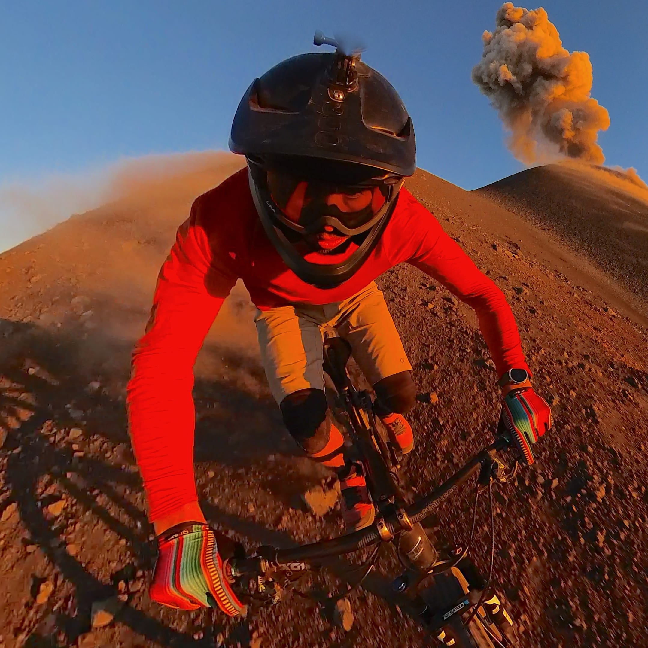 A cyclist in a Norrøna red shirt rides down a volcano, with a plume of smoke rising in the background and the sunrise to the right.