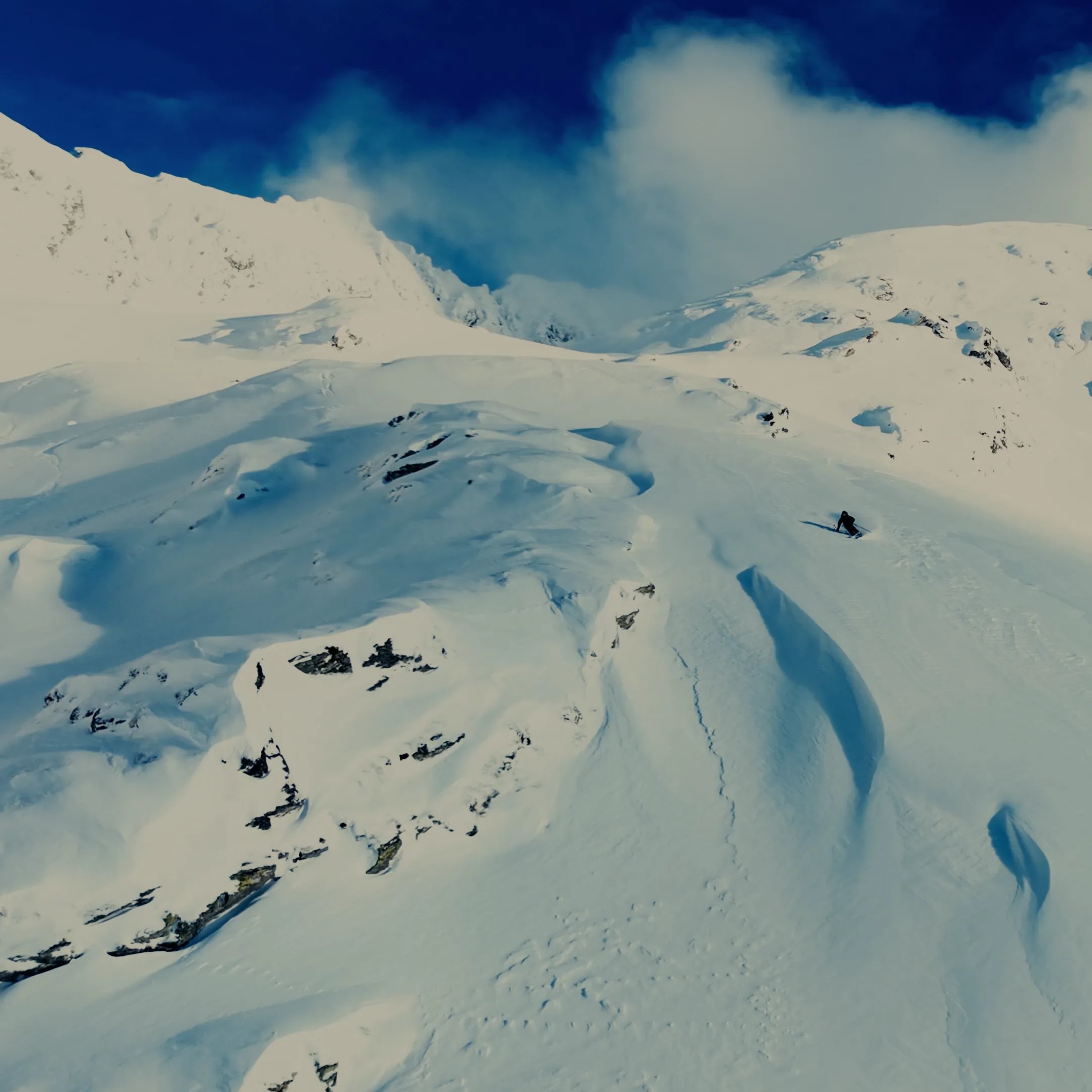 A skier in Norrøna outerwear descends a vast, snow-covered mountain slope.