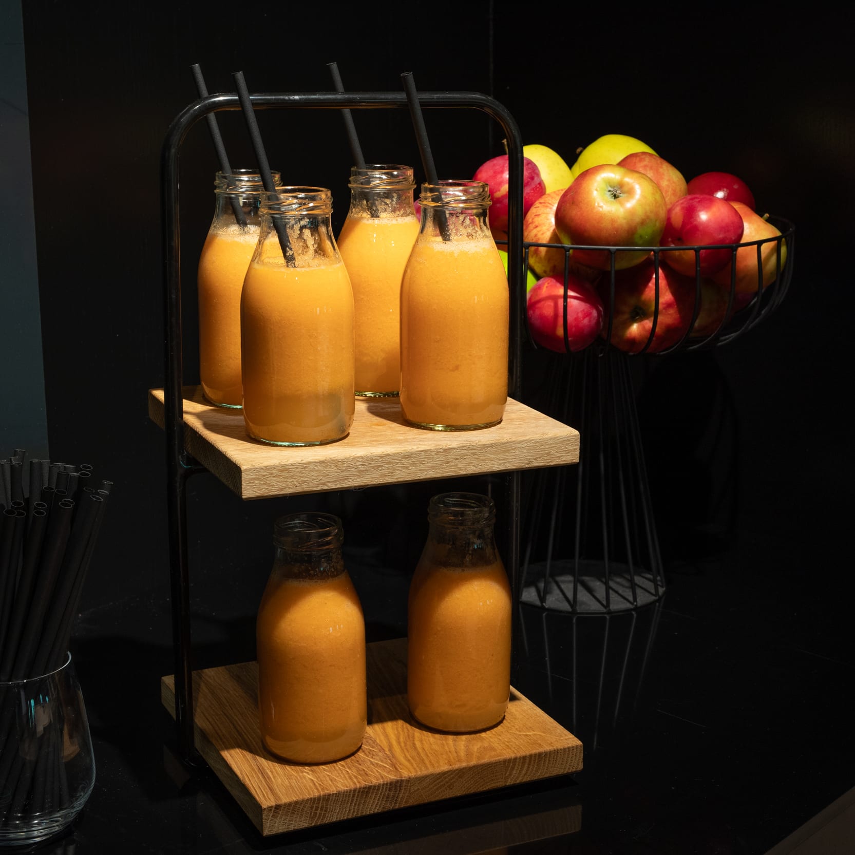 Bottles of orange juice with straws and a wire basket of apples are displayed on a black surface.