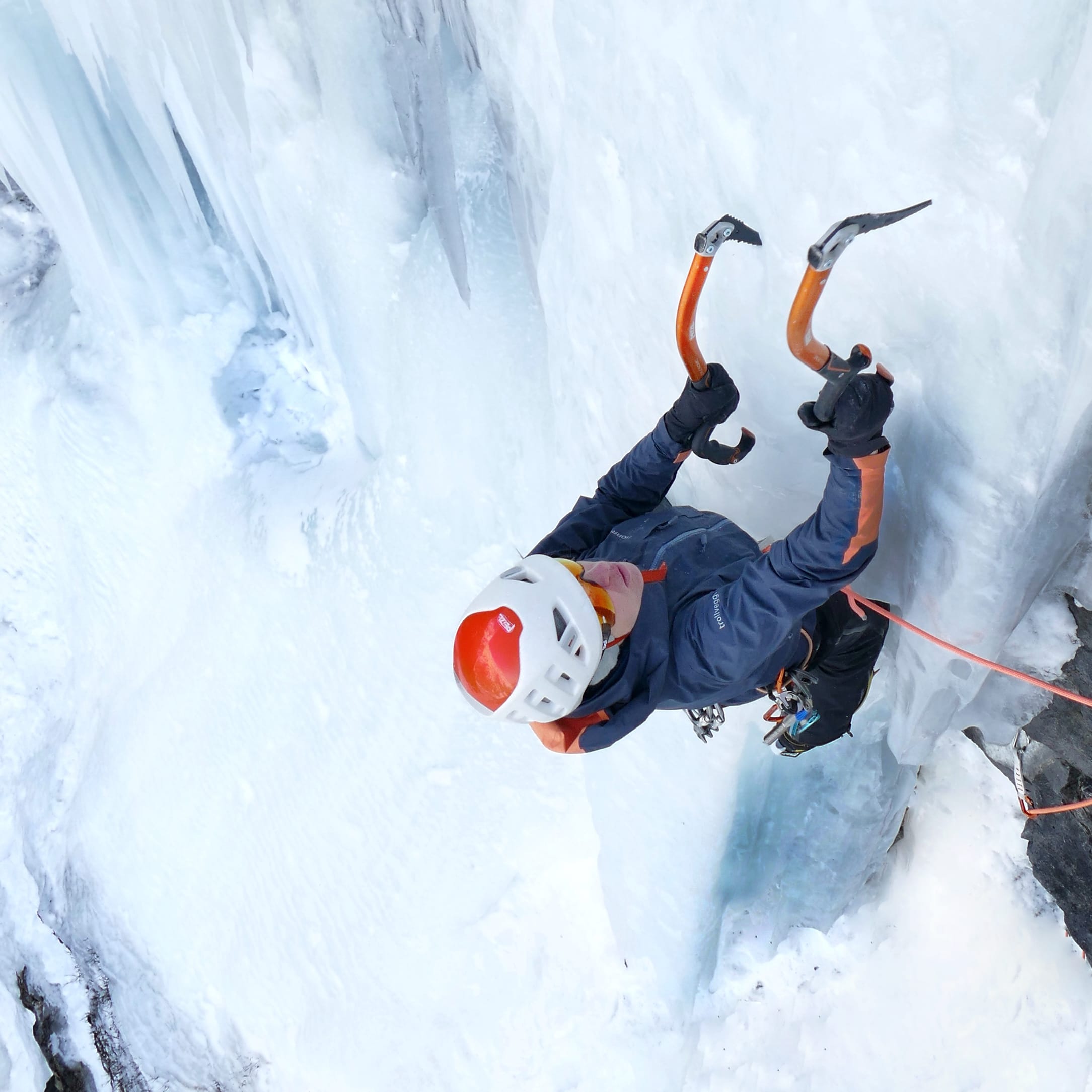 A person ice climbing a frozen waterfall, wearing a Norrøna jacket, helmet, gloves, and using ice axes.
