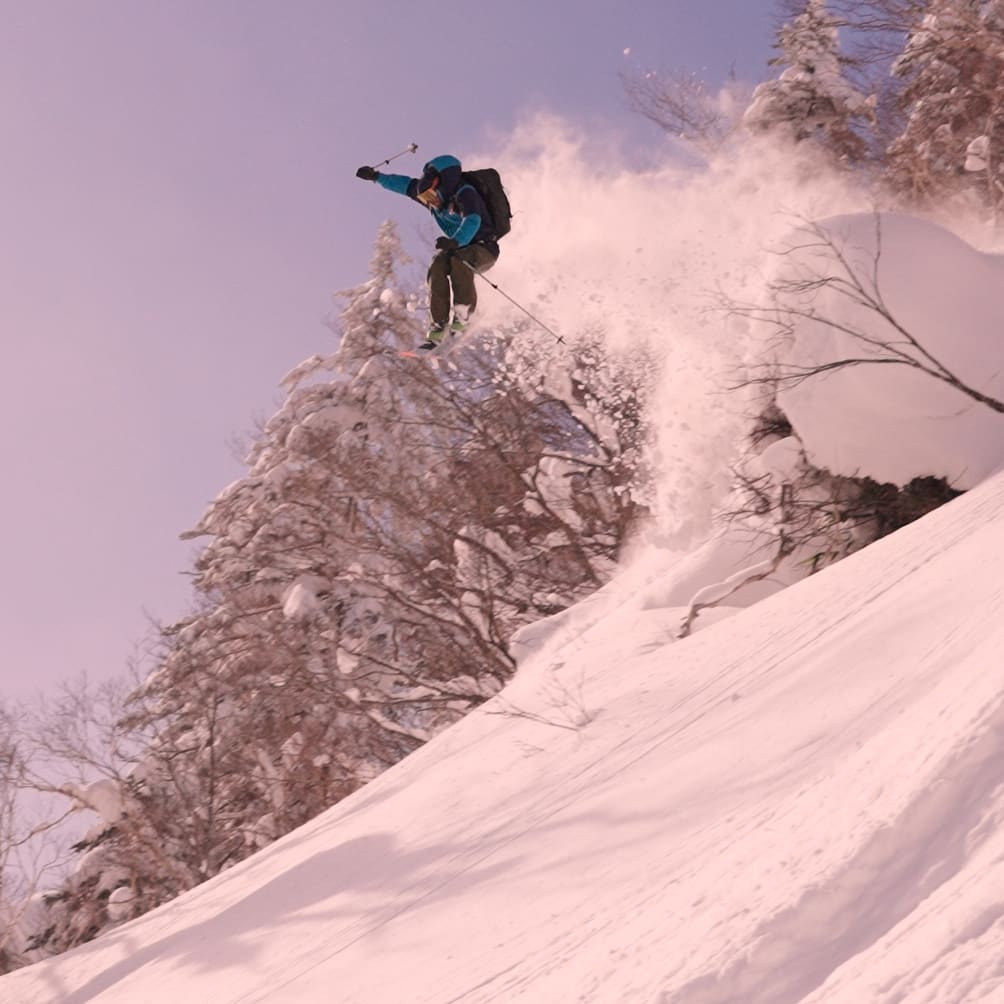 A skier in Norrøna gear jumps through snow covered trees, creating a powder cloud against a pale sky.