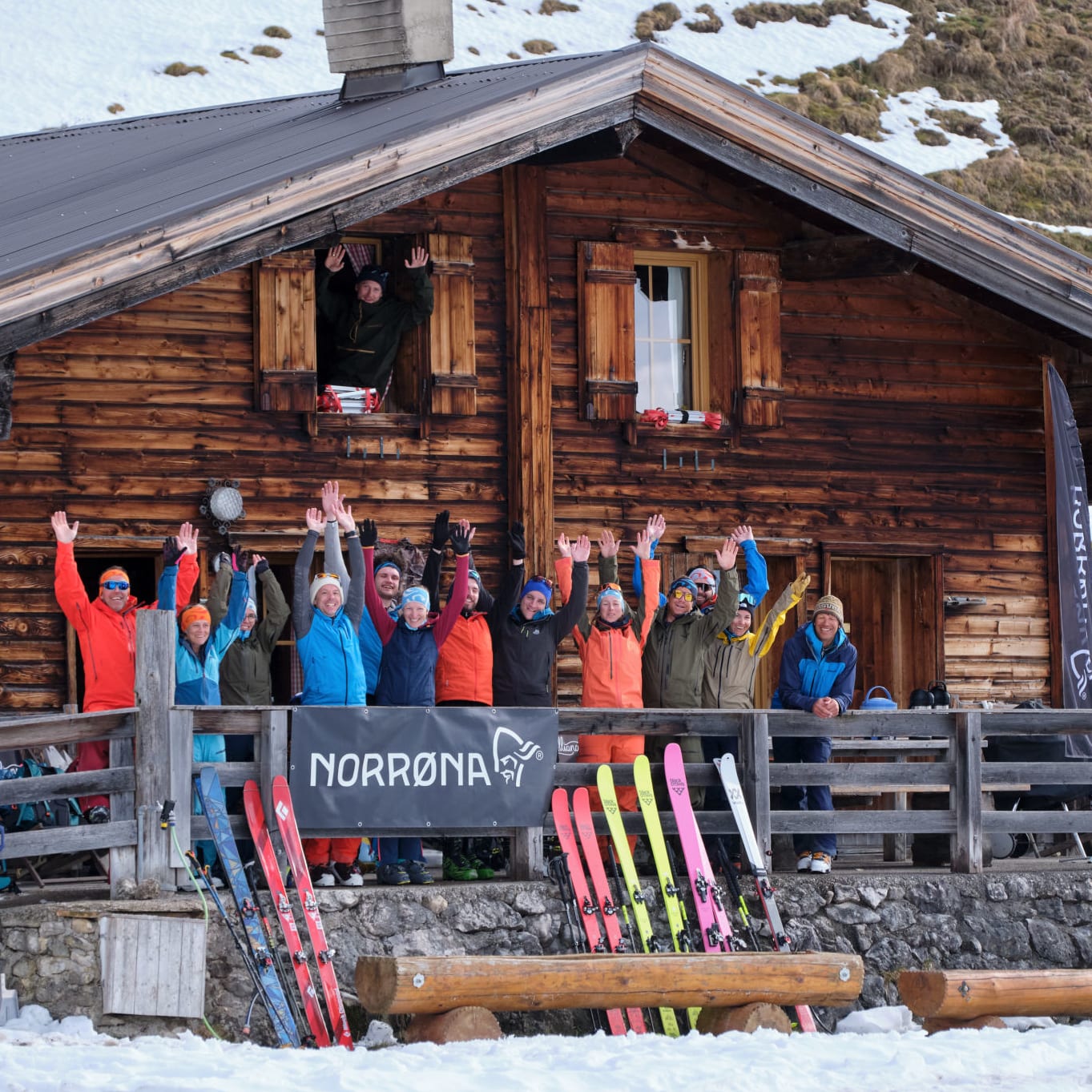 Group of people with skis outside a wooden building, waving with Norrøna flags and banner. Many wear Norrøna apparel. Snow surrounds the building.
