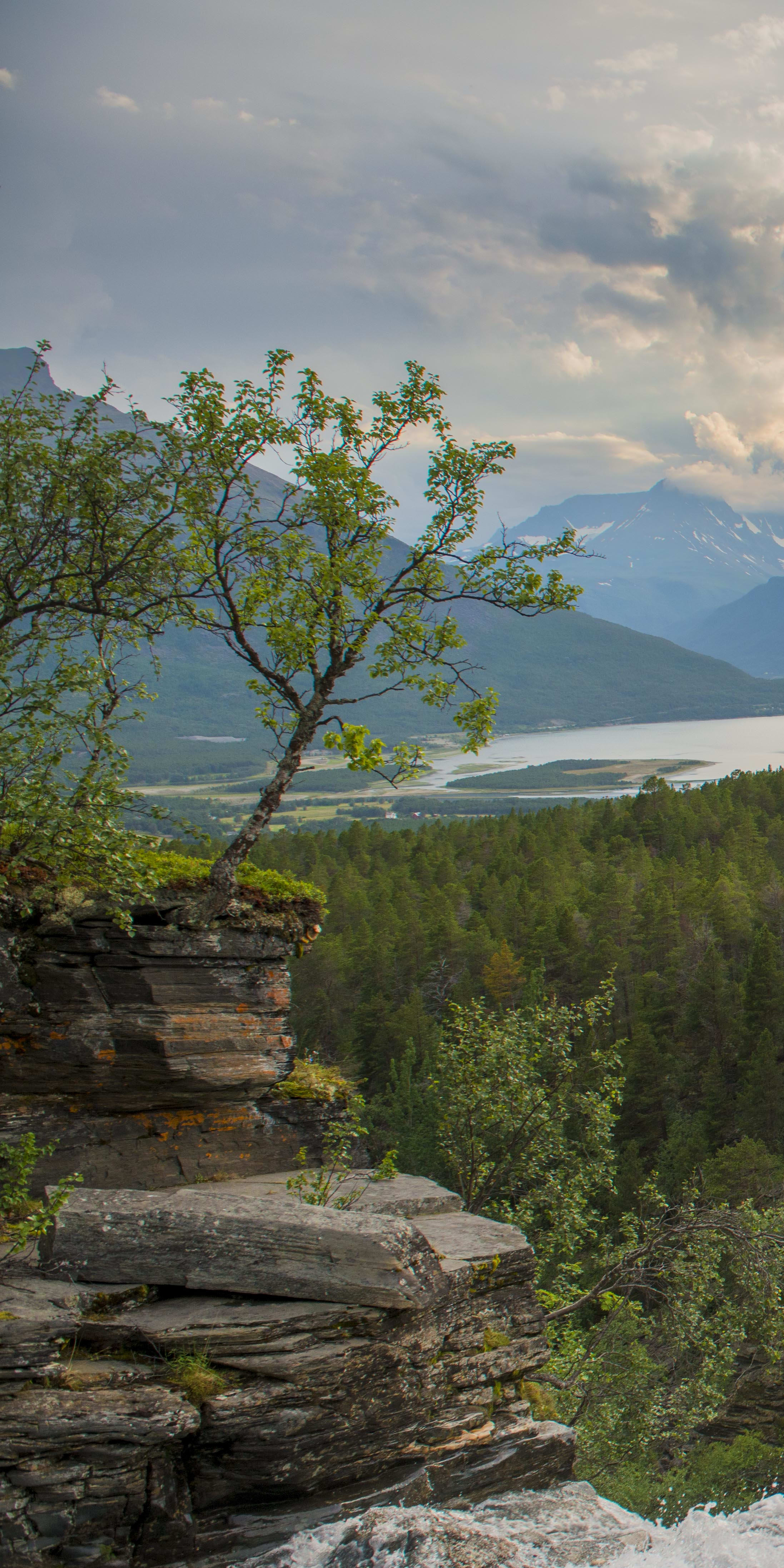 A view from a clifftop, overlooking a waterfall and a lake, with mountains in the background. 
